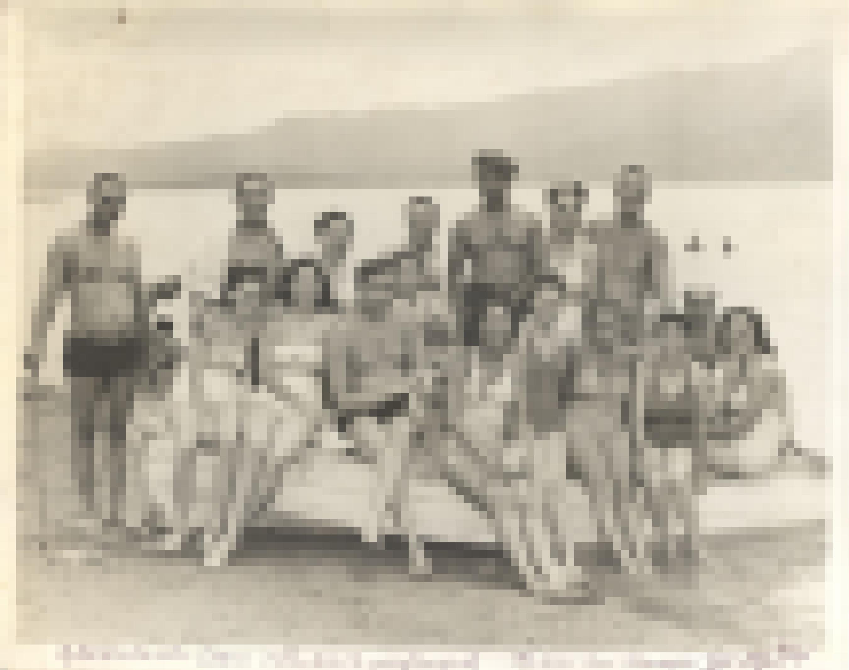 Eine Gruppe von etwa 20 Freundinnen und Freunden posiert in Badekleidung rund um ein Holzboot am See Lake Elsinore im Süden Kaliforniens. Sie lachen in die Kamera, hinter ihnen andere Badende im See, ca. 1940.