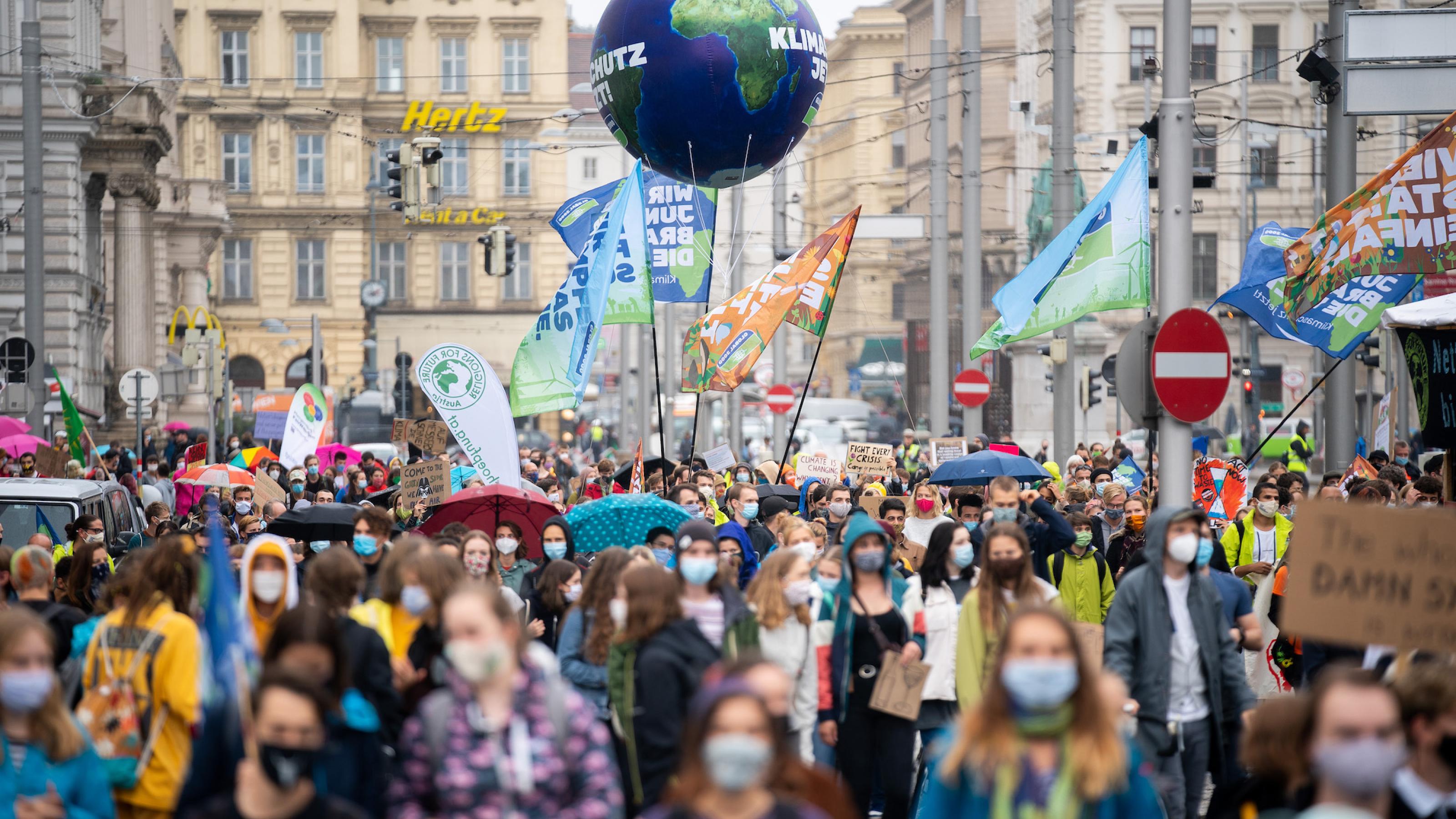 Dargestellt ist eine große Menschenmenge, die sich mit Bannern und Plakaten durch die Innenstadt von Wien bewegt. Auch eine aufblasbare Erdkugel haben die Demonstranten von Fridays for Future dabei. Im Gegensatz zu den Querdenkern tragen sie Gesichtsmasken.