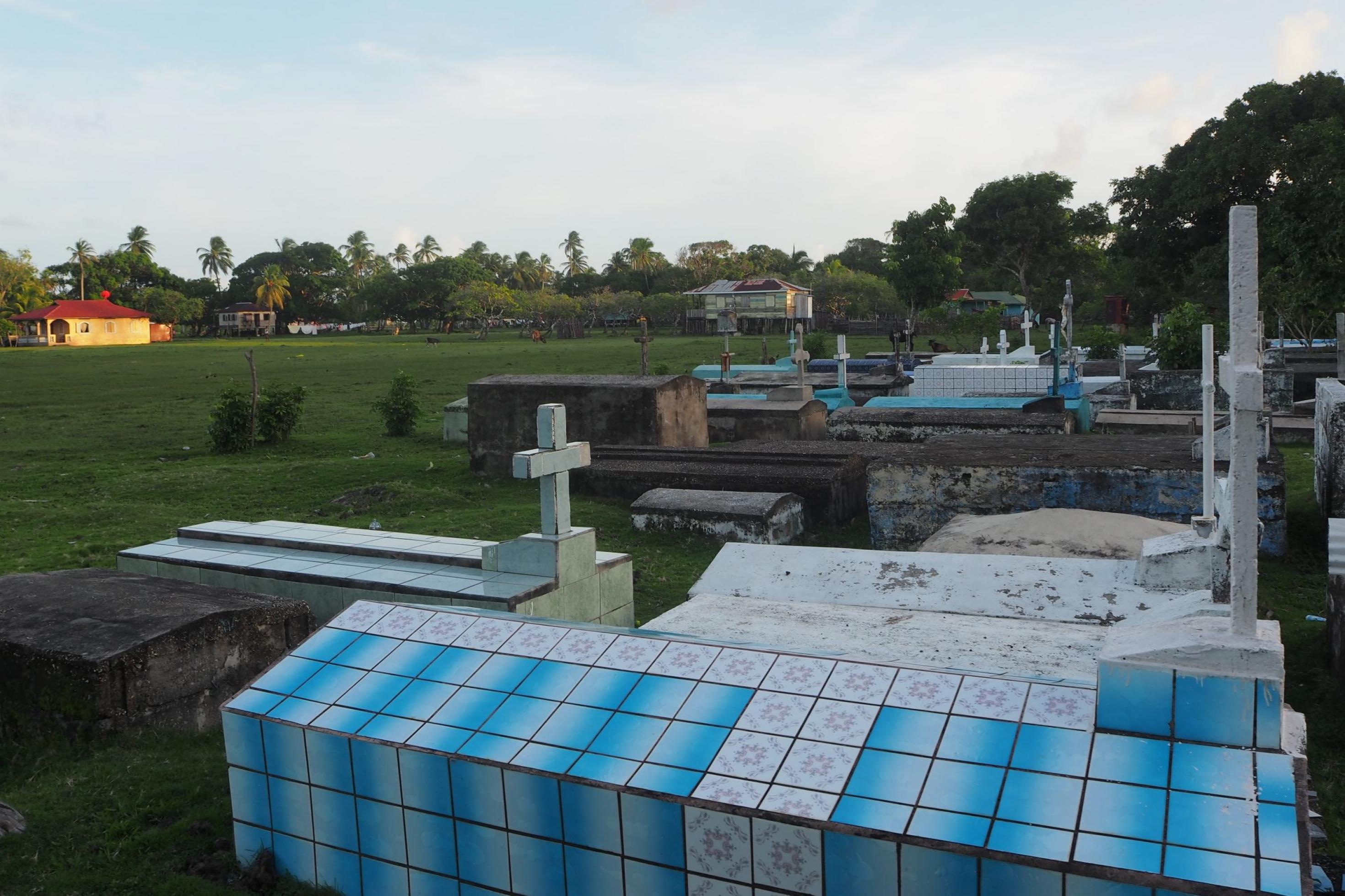 Weiß-blau gekachelte Gräber mit Betonkreuzen vor Palmen und Holzhäusern auf Pfählen