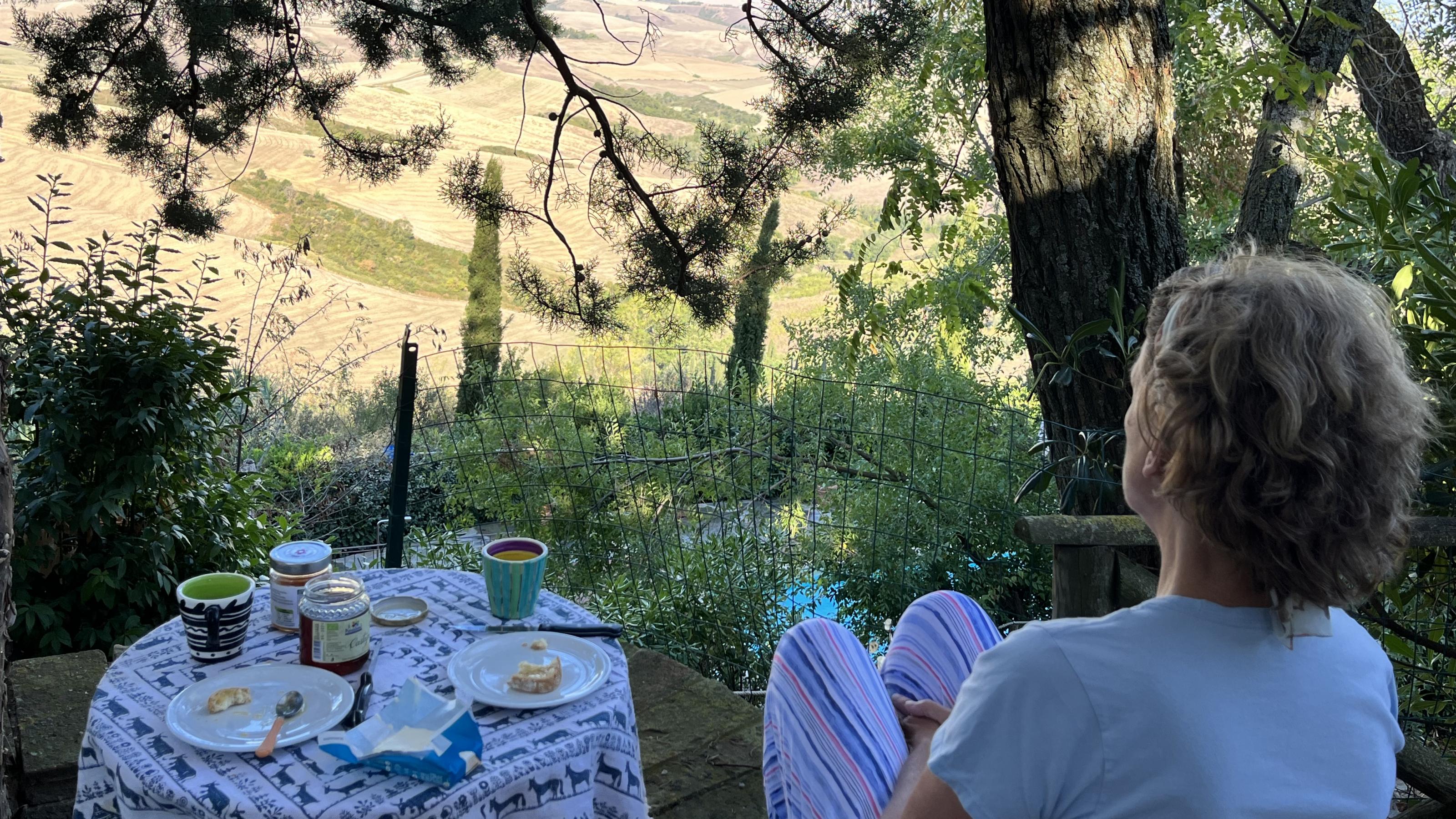 Eine Frau sitzt mit dem Rücken zum Betrachter neben einem kleinen runden Tisch, auf dem Frühstück angerichtet ist. Sie blickt vorbei an Ästen über gepflügte Felder der Toskana