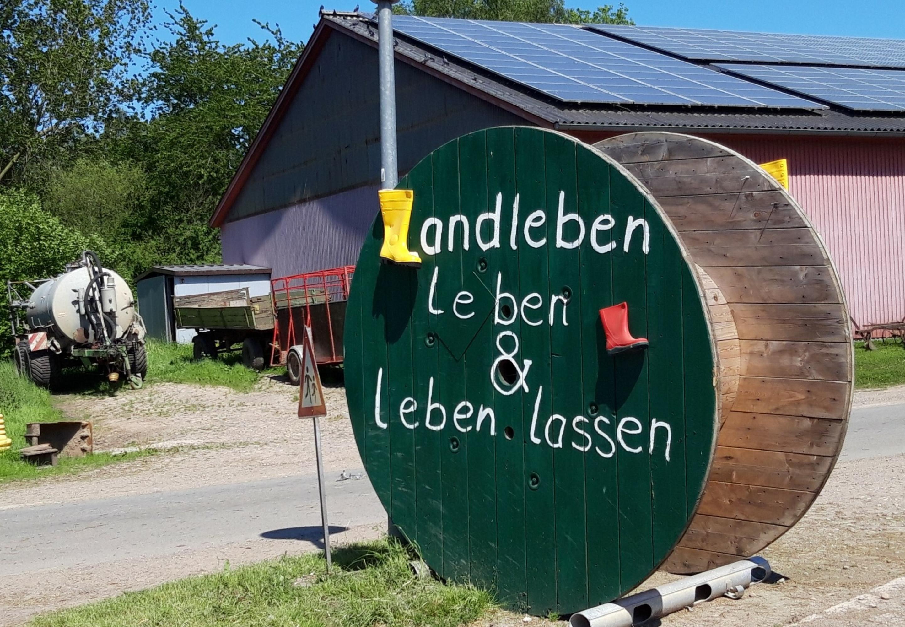 An der Straße ruht eine übermannshohe Schlauchtrommel. Sie ist grün gestrichen, mit alten farbigen Kinderstiefeln dekoriert und trägt die Aufschrift „Landleben – leben & leben lassen“.