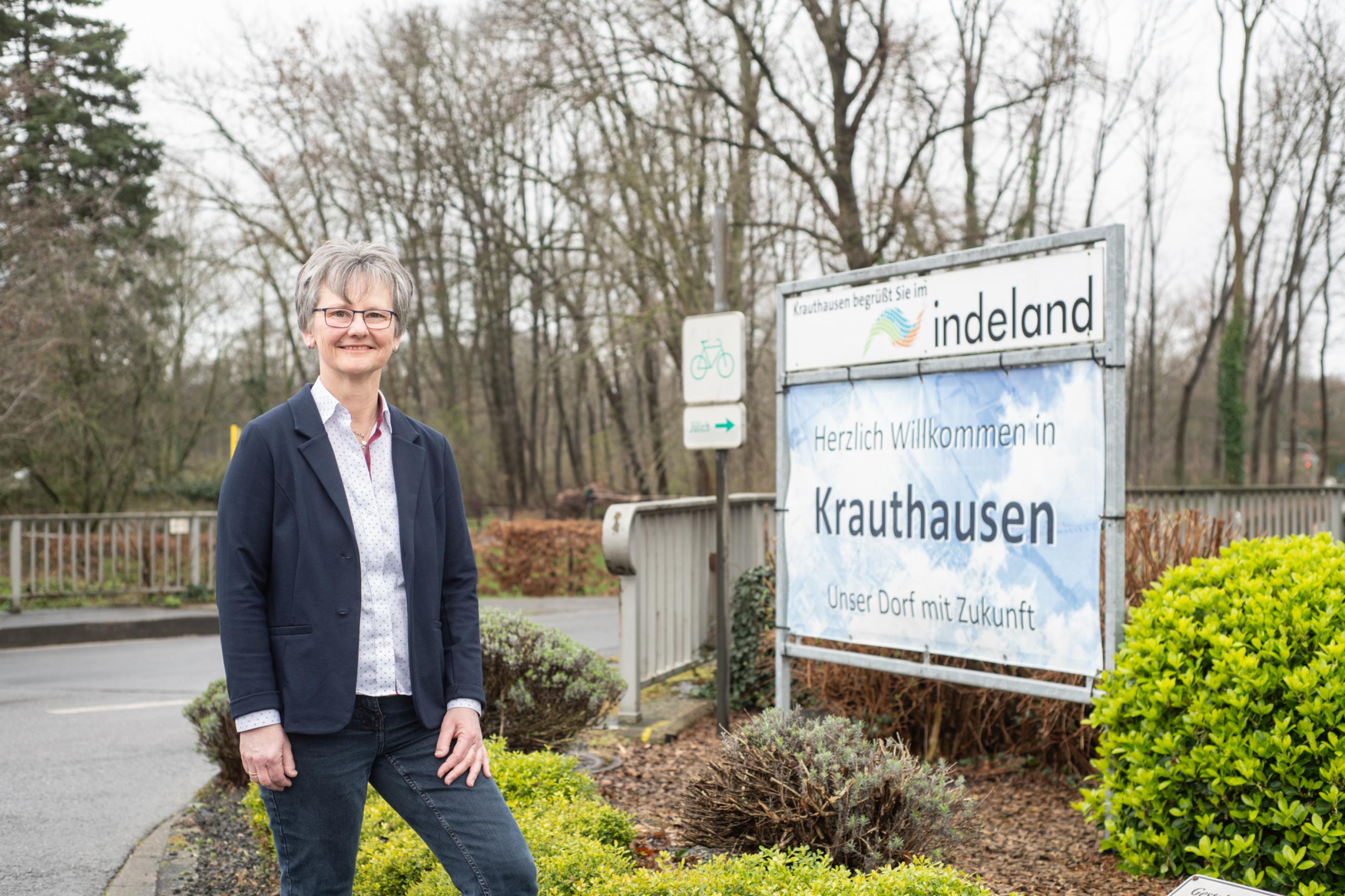 Eine Frau mit grauen Haaren, Brille und Alltagskleidung steht auf einem bepflanzten Kreisverkehr mit einem Schild, auf dem „Krauthausen“ steht.