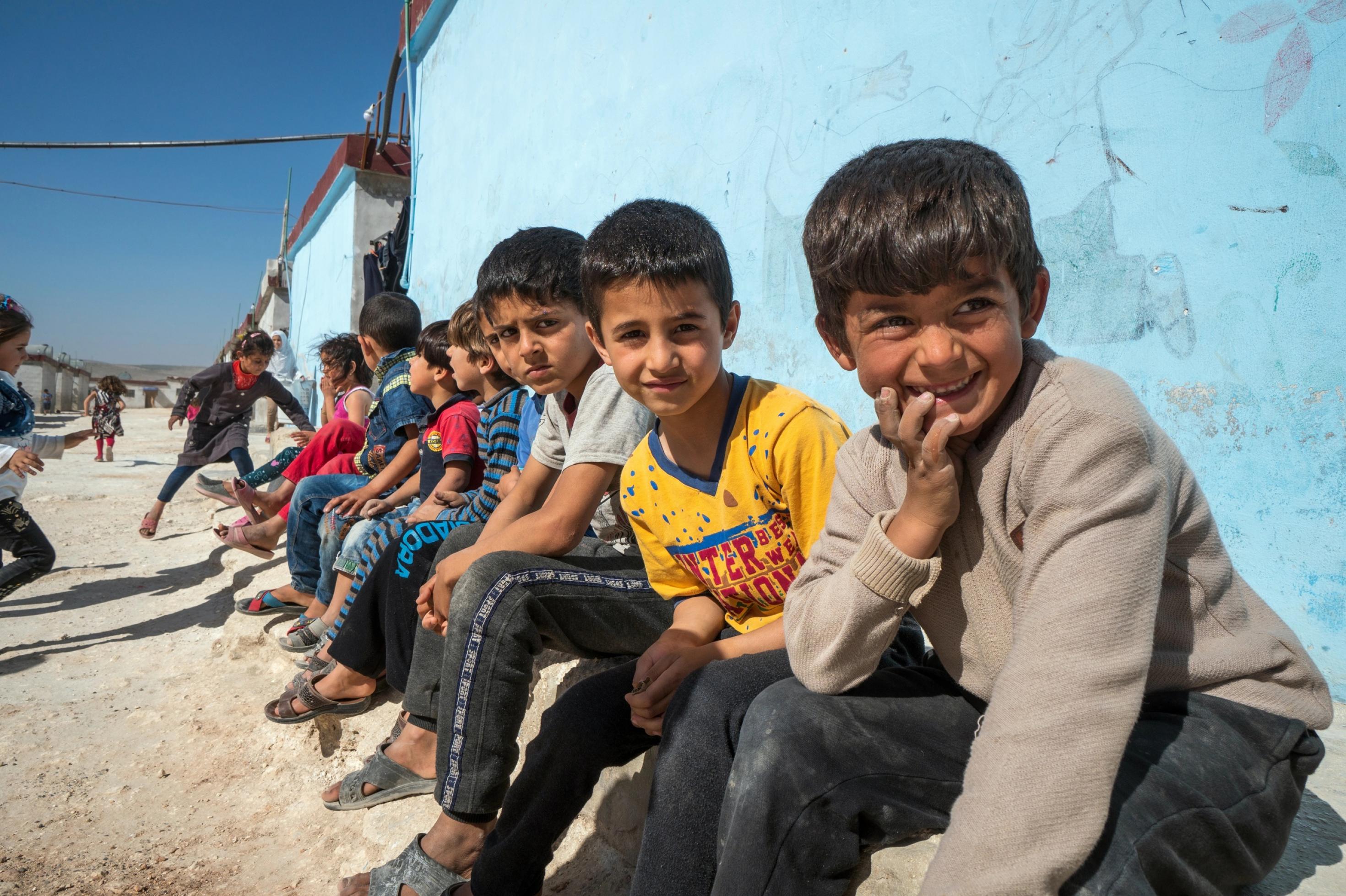 Eine Handvoll Kinder sitzen nebeneinander aufgereiht auf einer Bank vor einer hellblauen Hauswand. Im Vordergrund drei Jungen, die eher schwermütig in die Kamera blicken.