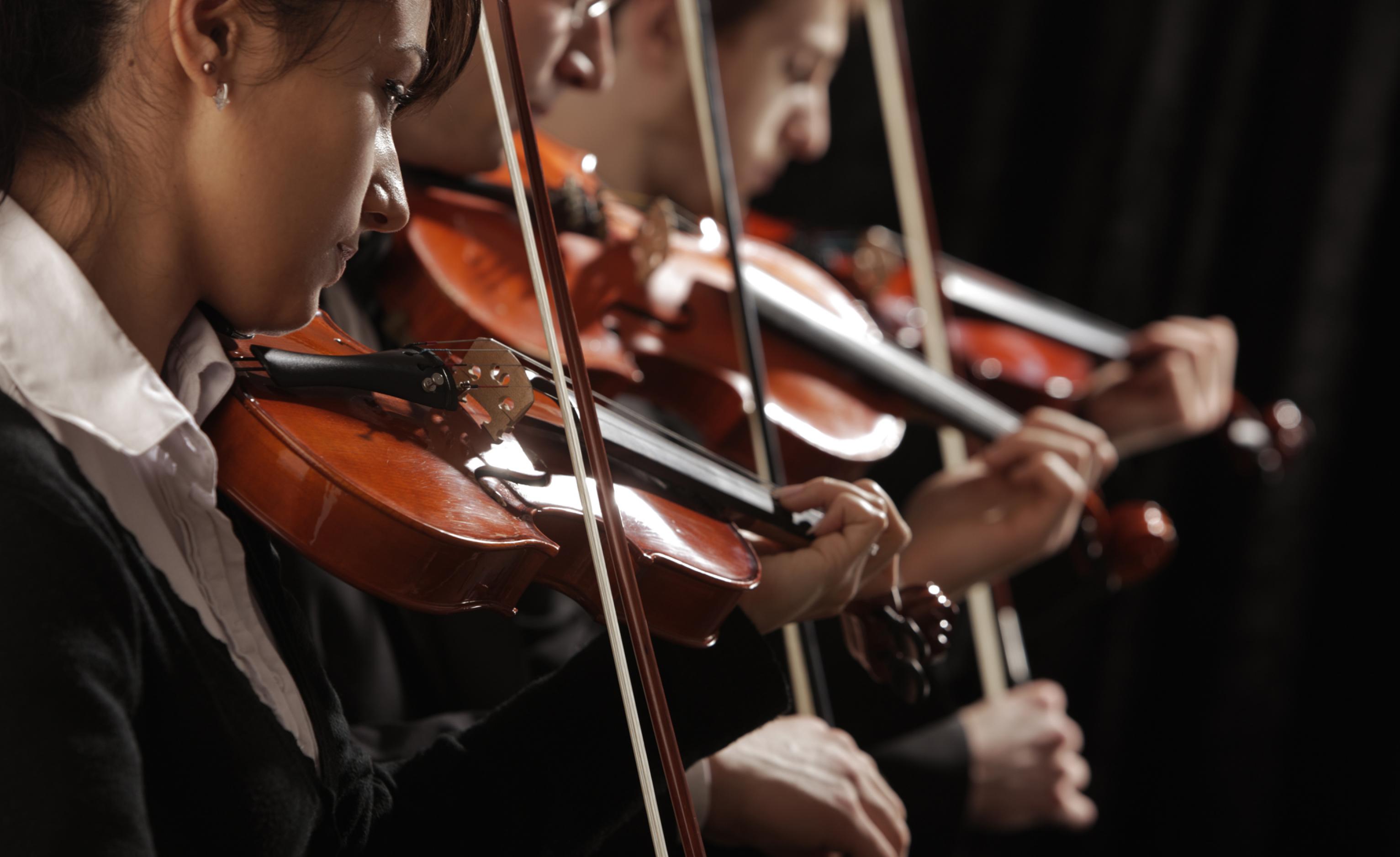 Das Foto zeigt vor schwarzem Hintergrund drei Geigende eines Orchesters. Sie sind elegant gekleidet und von der Seite fotografiert. Kopf, Oberkörper, der rechte Arm mit dem Geigenbogen und die Geigen selbst sind groß zu sehen.