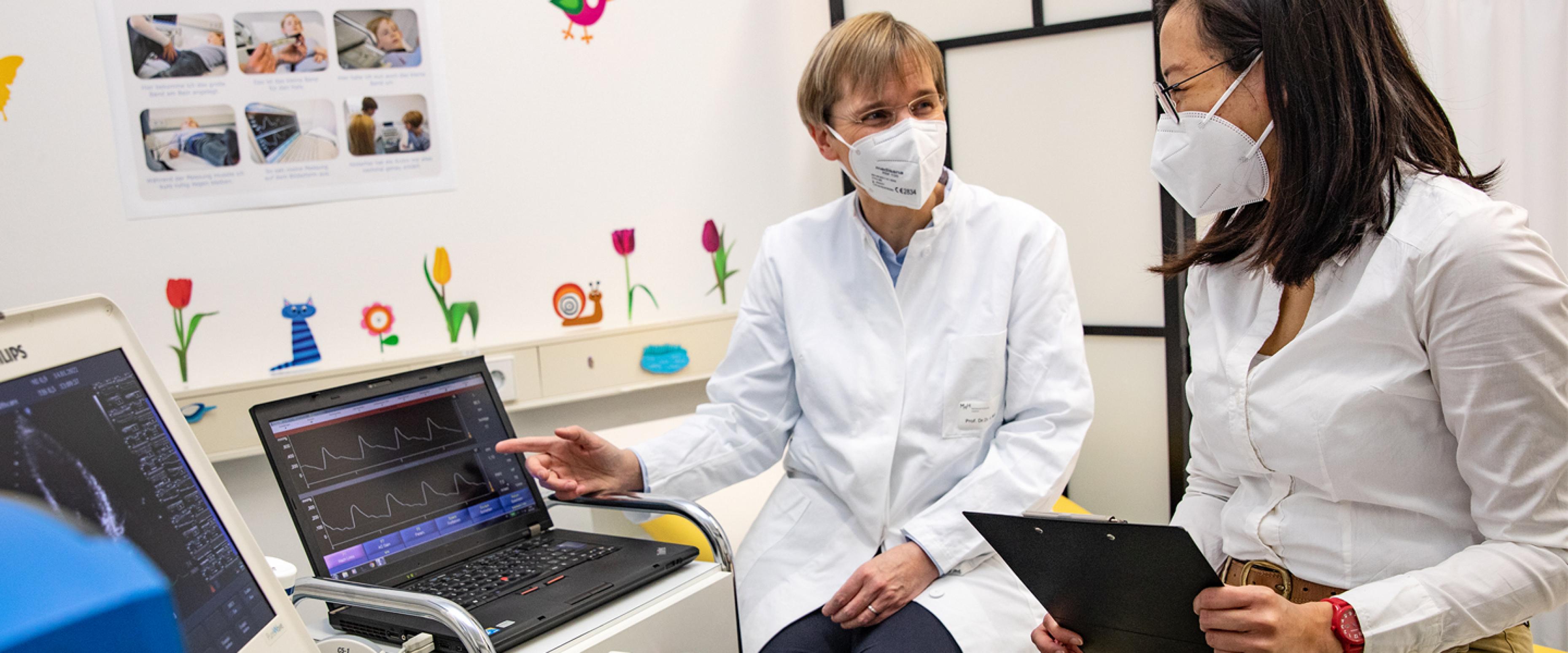 Das Bild zeigt zwei Frauen mit weißem Kittel und FFP2-Maske, die Messergebnisse auf einem Computer besprechen.