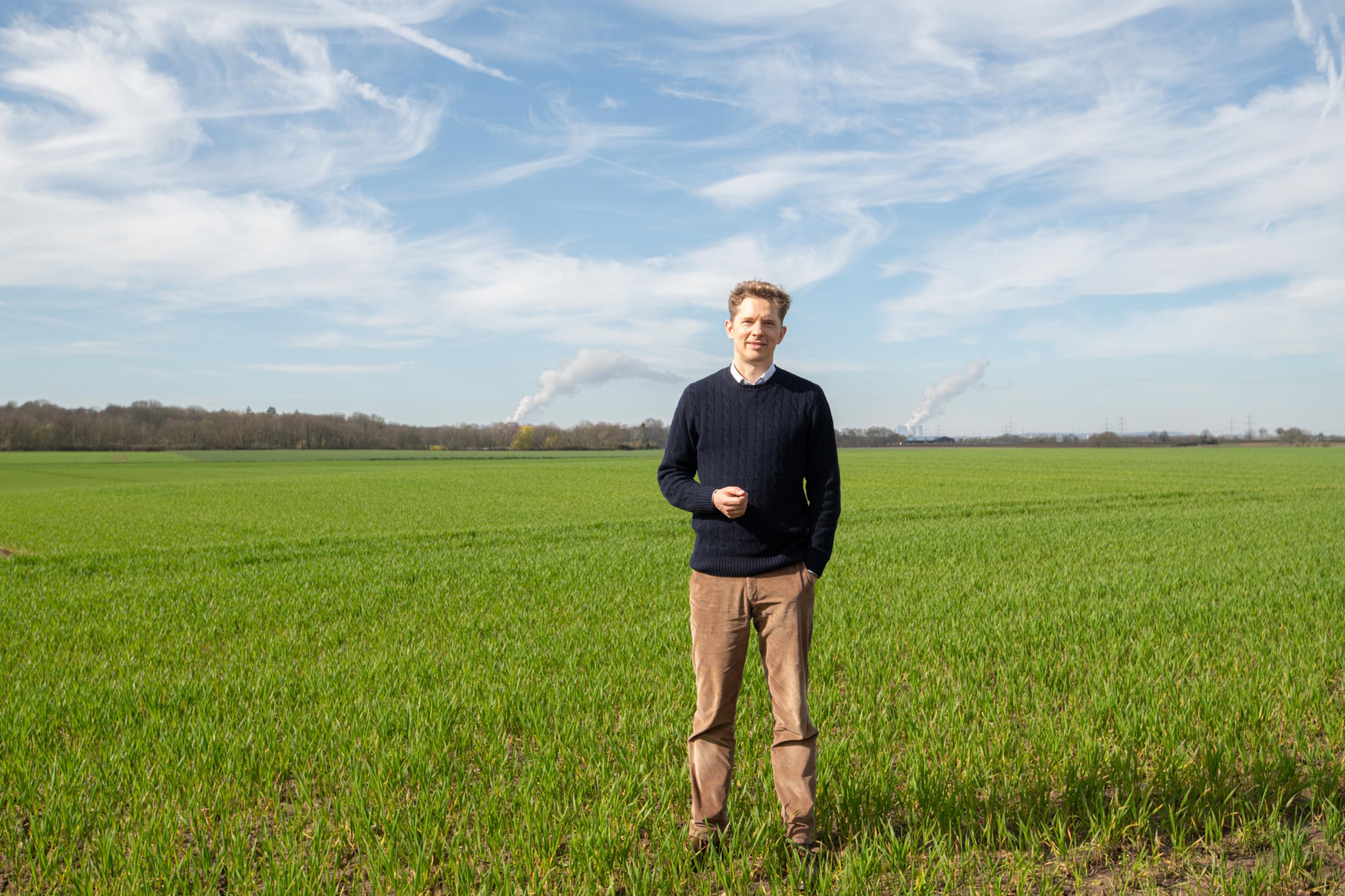 Ein junger Mann in brauner Hose und blauem Pullover steht auf einem Acker.