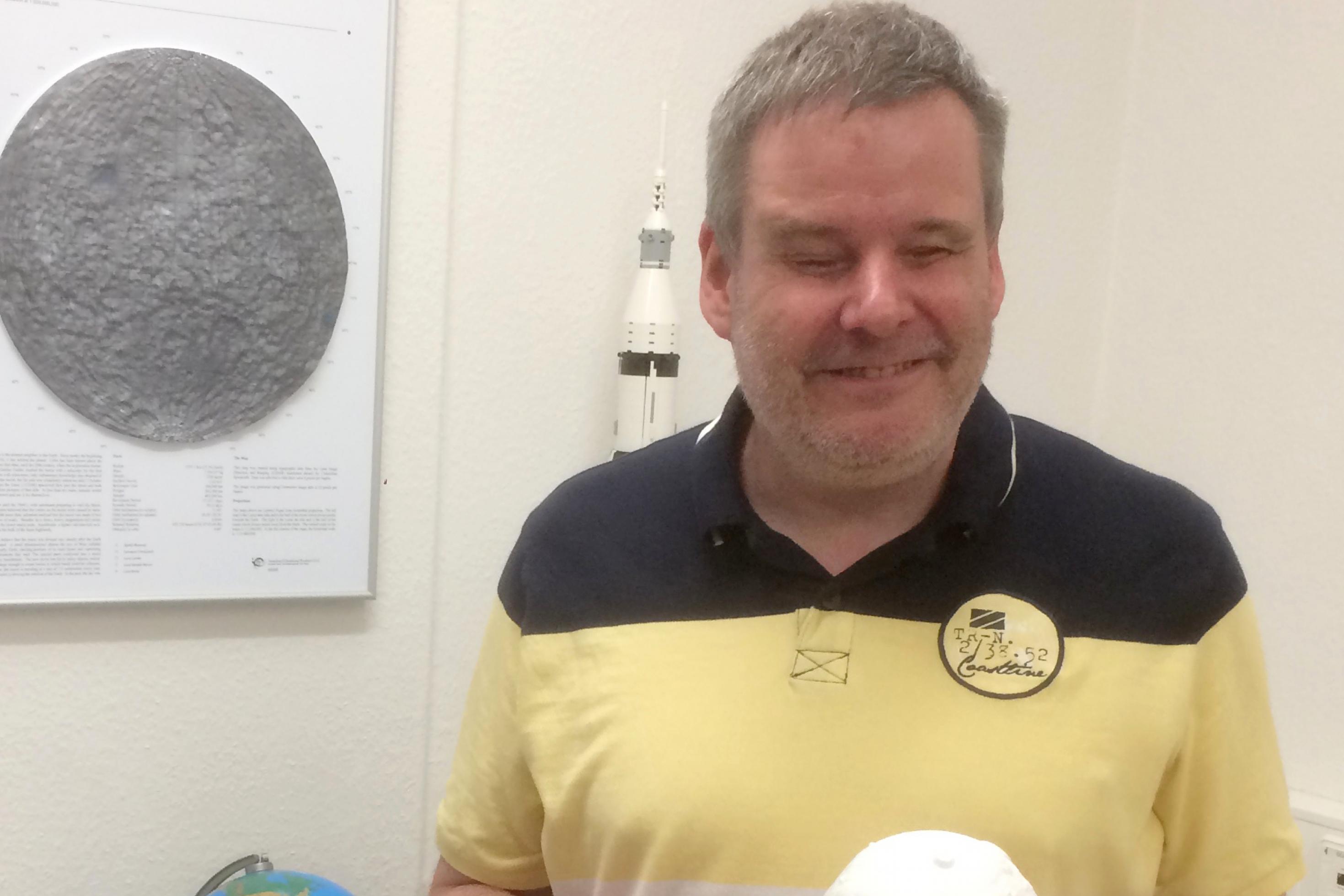 Gerhard Jaworek hält eine weiße Kugel (ein 3D-gedruckter Planet), hinter ihm liegen auf einem Tisch allerlei Globen und der Komet 67P als Modell