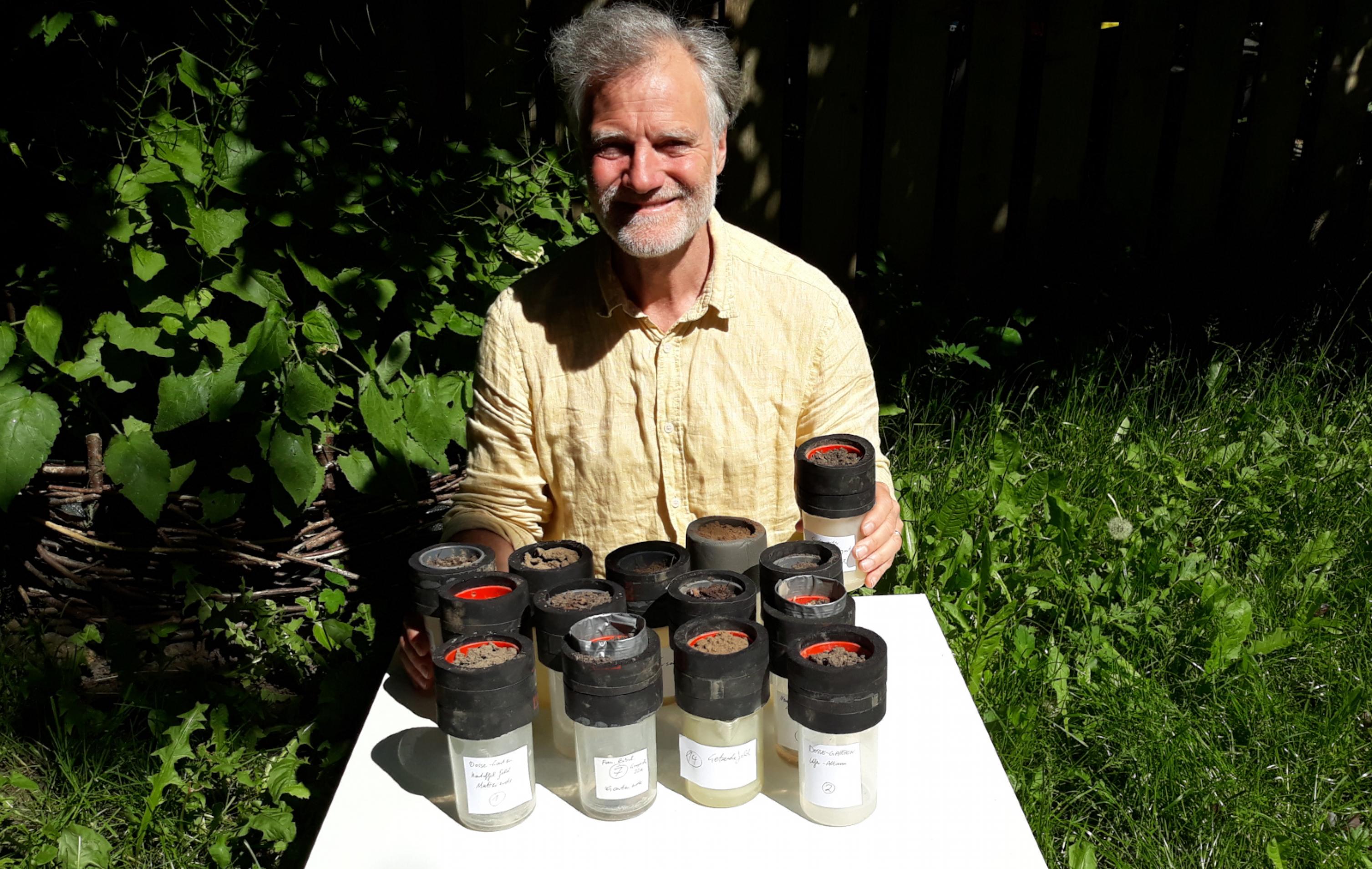 Ein Mann im Freien vor einem Klapptisch, darauf 14 Probenbehälter mit Bodenproben in verschiedensten Brauntönen