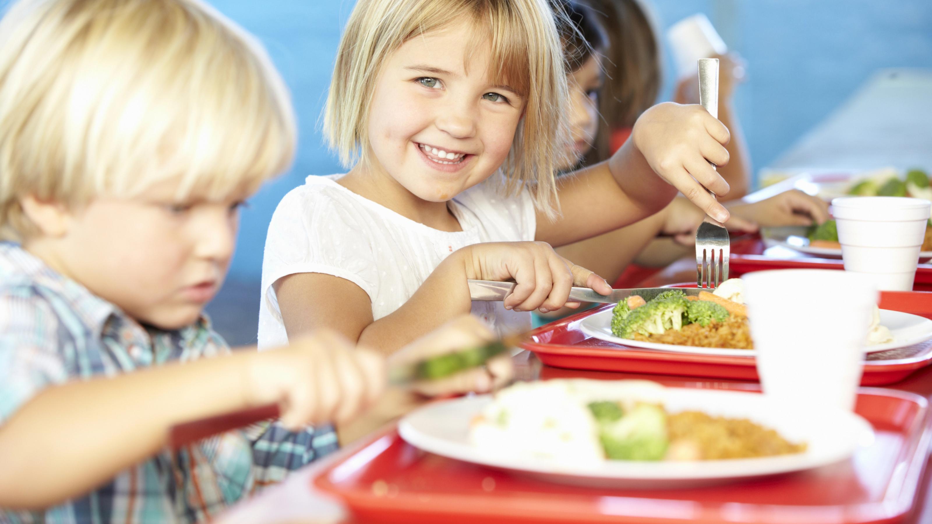 Fröhliche Kinder beim Essen in der Mensa, vor ihnen ein Teller unter anderem mit Brokkoli.