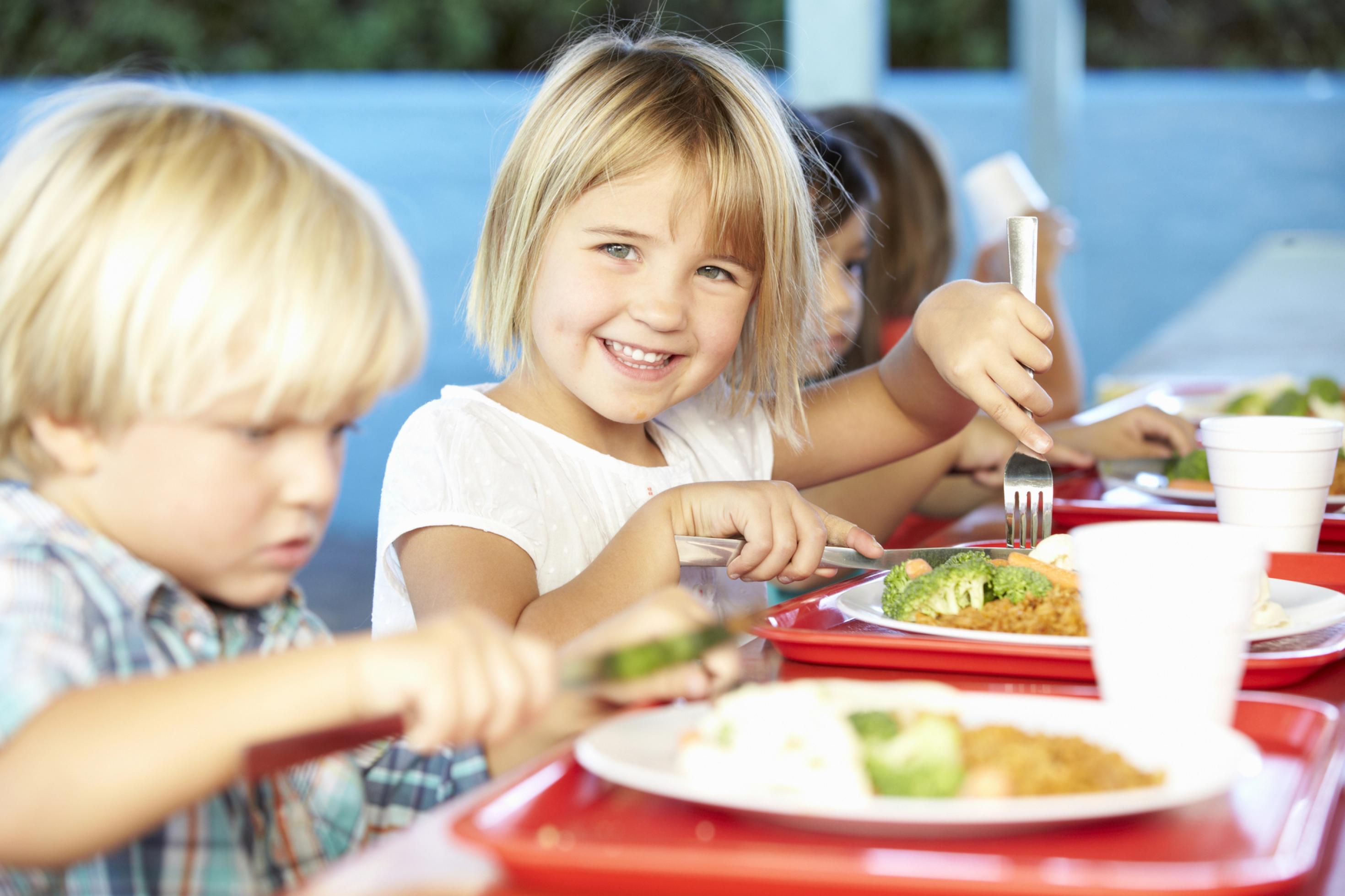 Fröhliche Kinder beim Essen in der Mensa, vor ihnen ein Teller unter anderem mit Brokkoli.