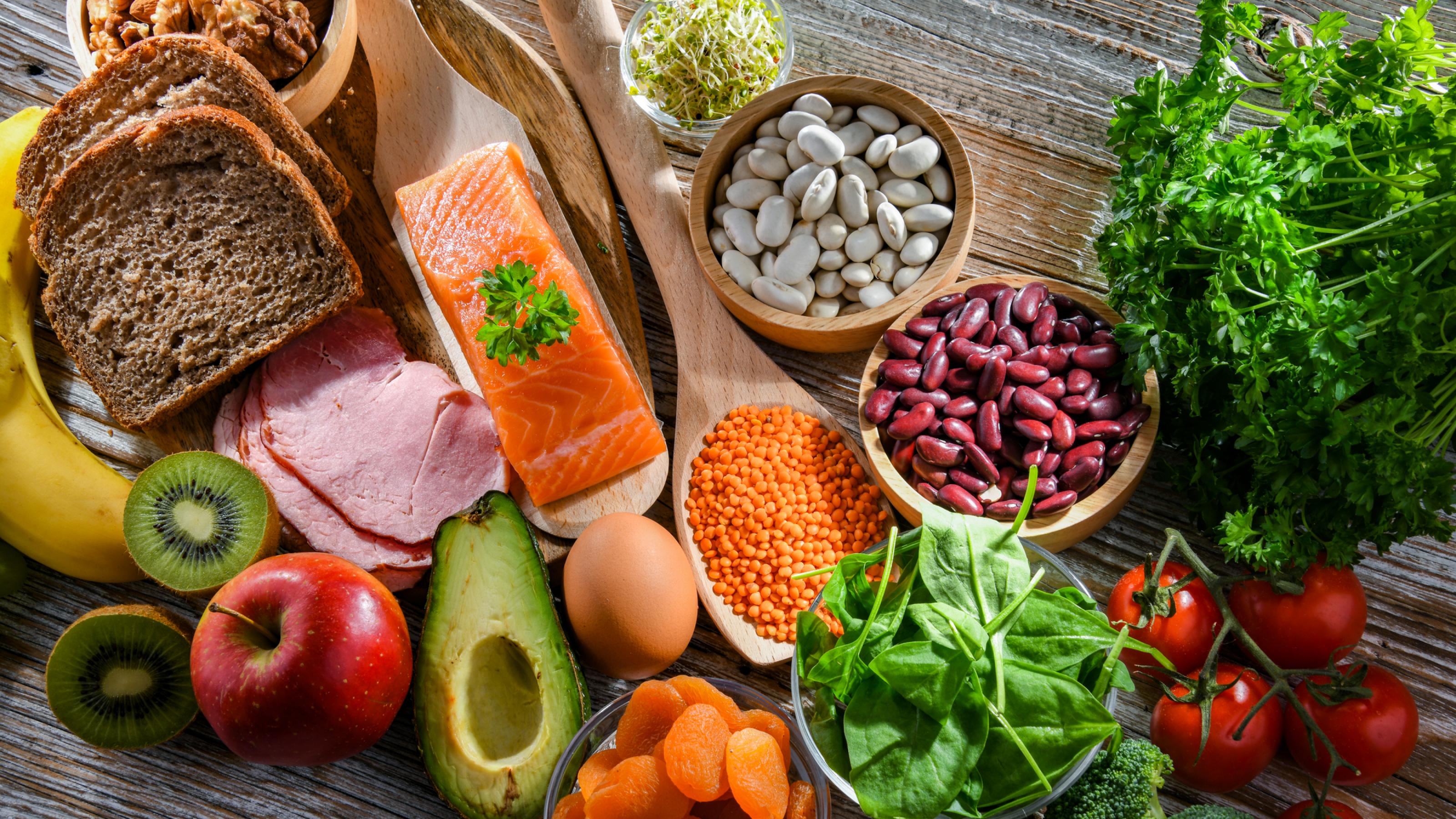 Draufsicht: Ein Überblick von gesunden Lebensmitteln, die auf einer Tischplatte liegen. Avocado, Vollkornbrot, Kräuter, Spinat, Tomaten, Brokkoli, Apfel, Kiwi, Lachs, Ei, Nüsse und Hülsenfrüchte.