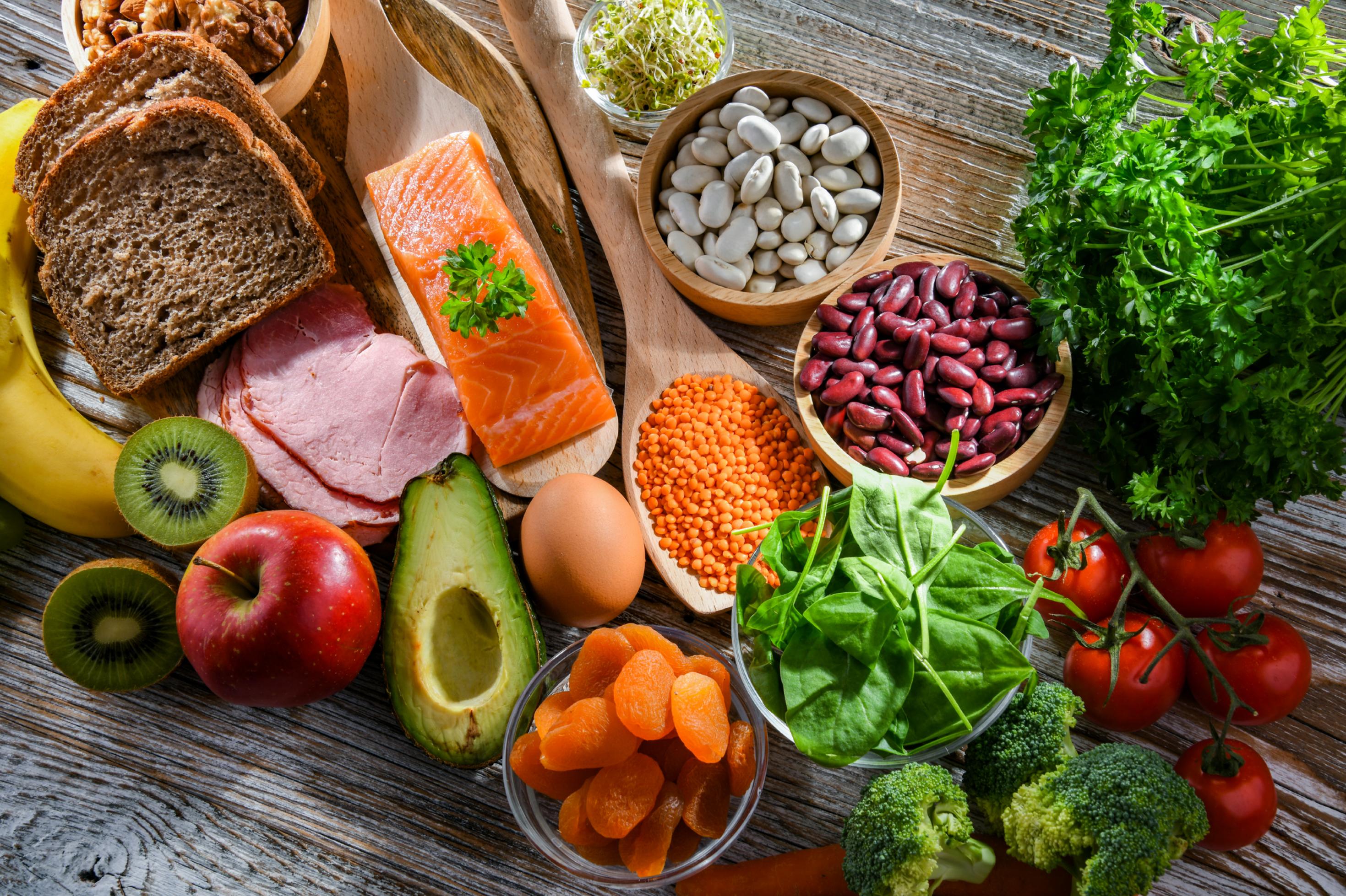 Draufsicht: Ein Überblick von gesunden Lebensmitteln, die auf einer Tischplatte liegen. Avocado, Vollkornbrot, Kräuter, Spinat, Tomaten, Brokkoli, Apfel, Kiwi, Lachs, Ei, Nüsse und Hülsenfrüchte.