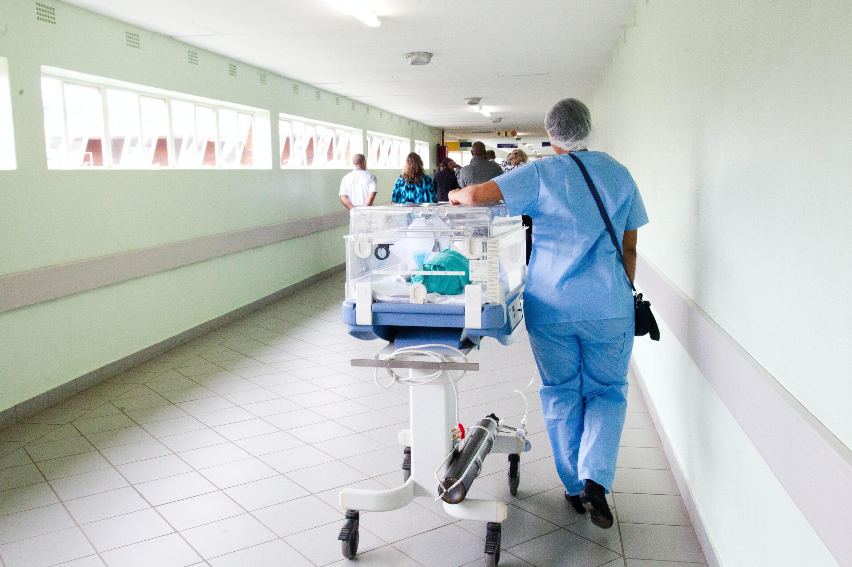 Ein Krankenhausflur, eine Krankenpflegerin schiebt einen Brutkasten. Im Hintergrund Besucher des Krankenhauses.
