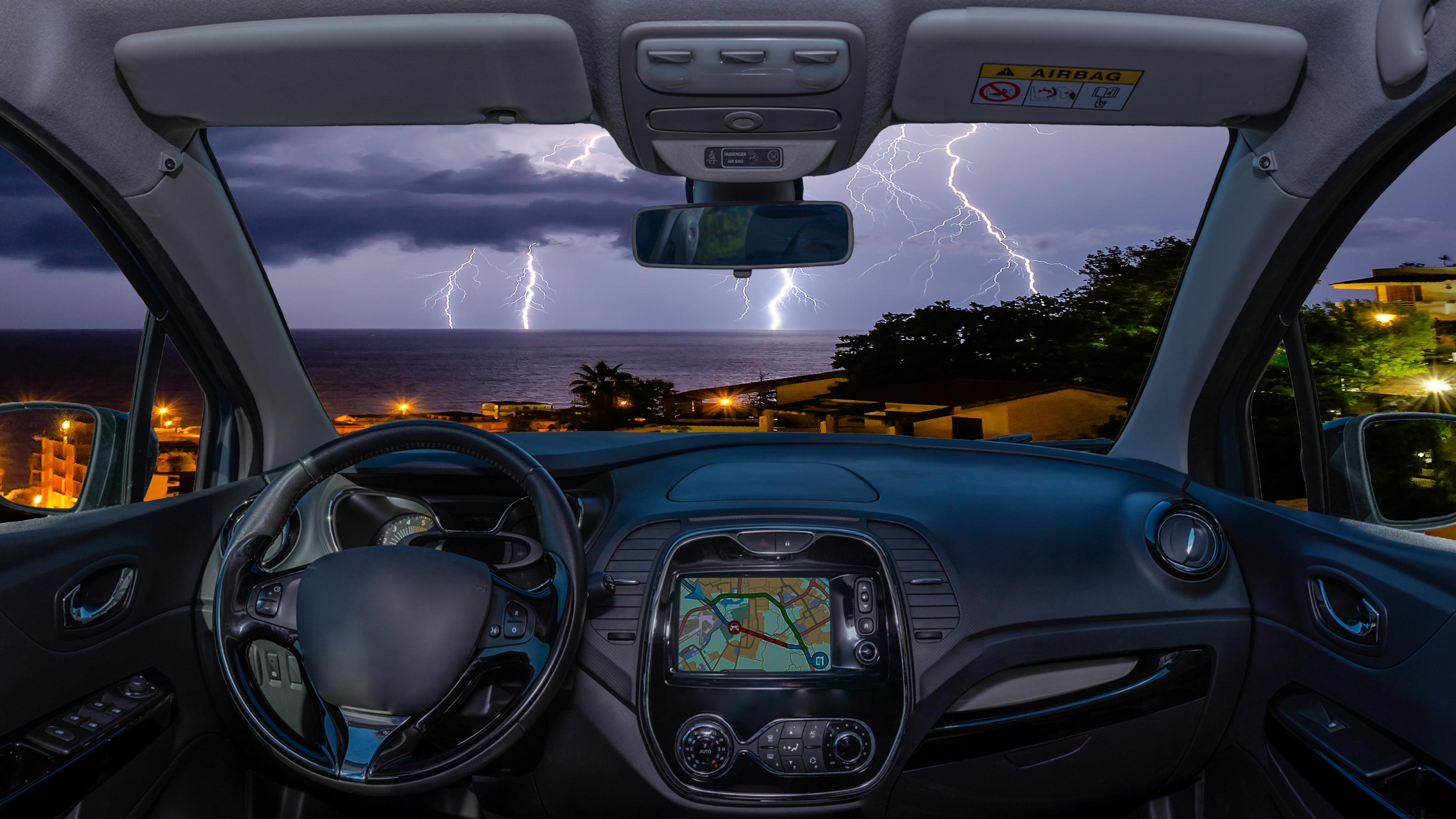 Blick über das Armaturenbrett eines Autos. Draußen sind mehrere Blitze zu sehen.