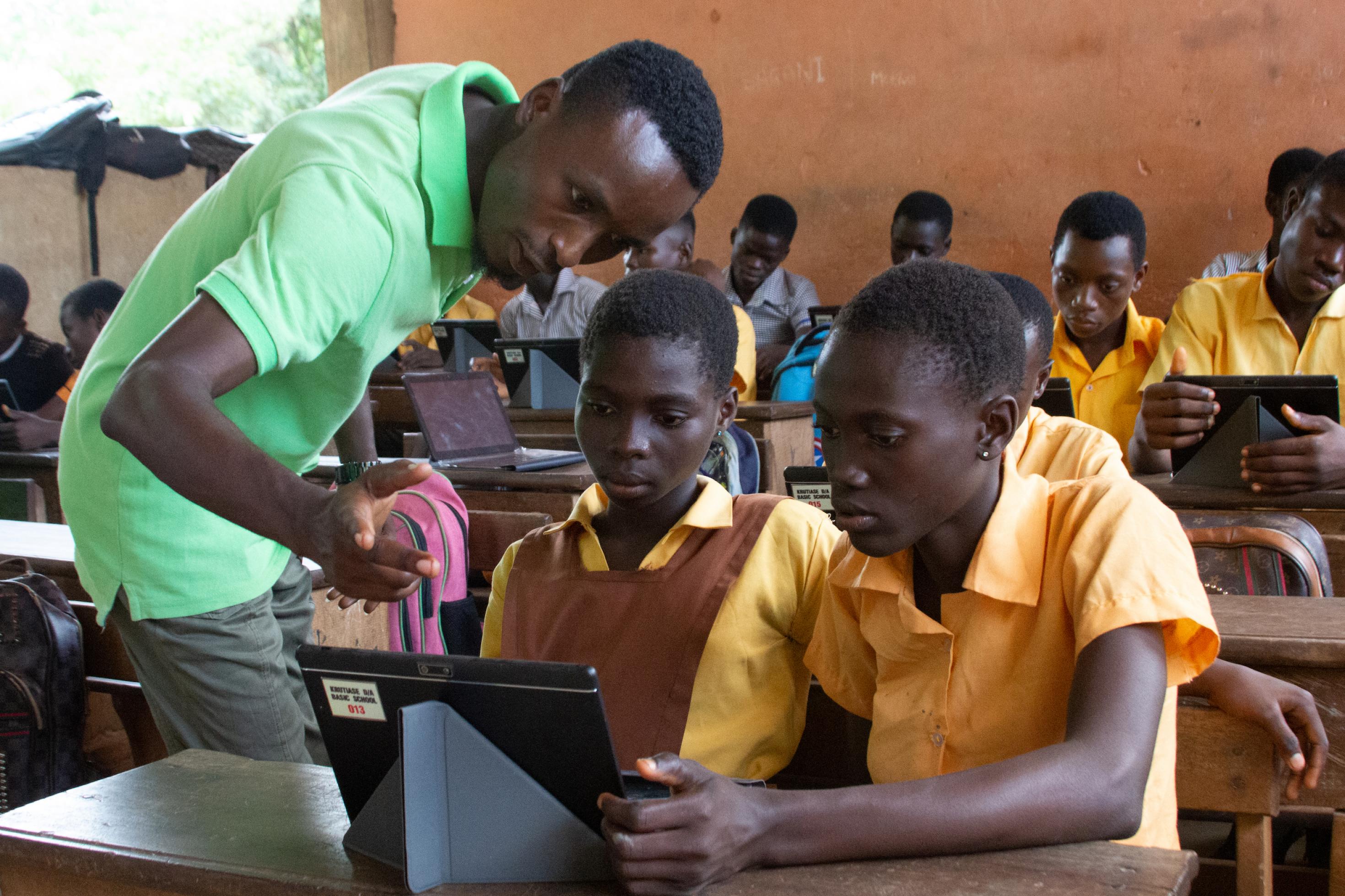 Ein junger Lehrer, leuchtend grünes T-Shirt, beugt sich über das Tablett. Zwei Schülerinnen, die an der Bank sitzen, folgen seinem Blick. Alle drei schauen in das Tablett.