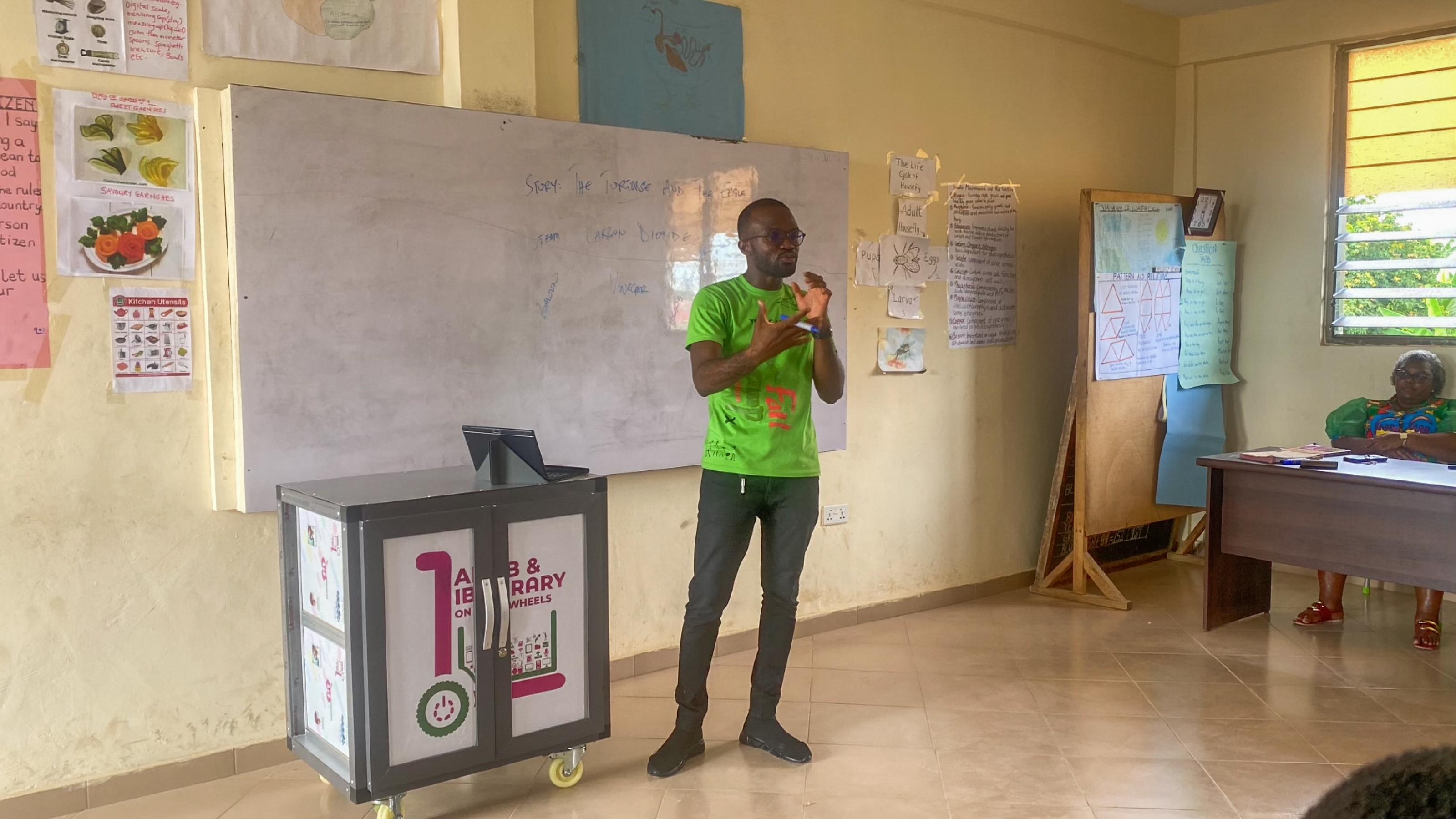 Ein Mann im hellgrünen T-Shirt vor einer Schulklasse, vor einem großen Whiteboard, neben sich das mobile Computerlabor – also ein Kasten mit Rädern, darauf die Aufschrift: Lab and Library.Wagen