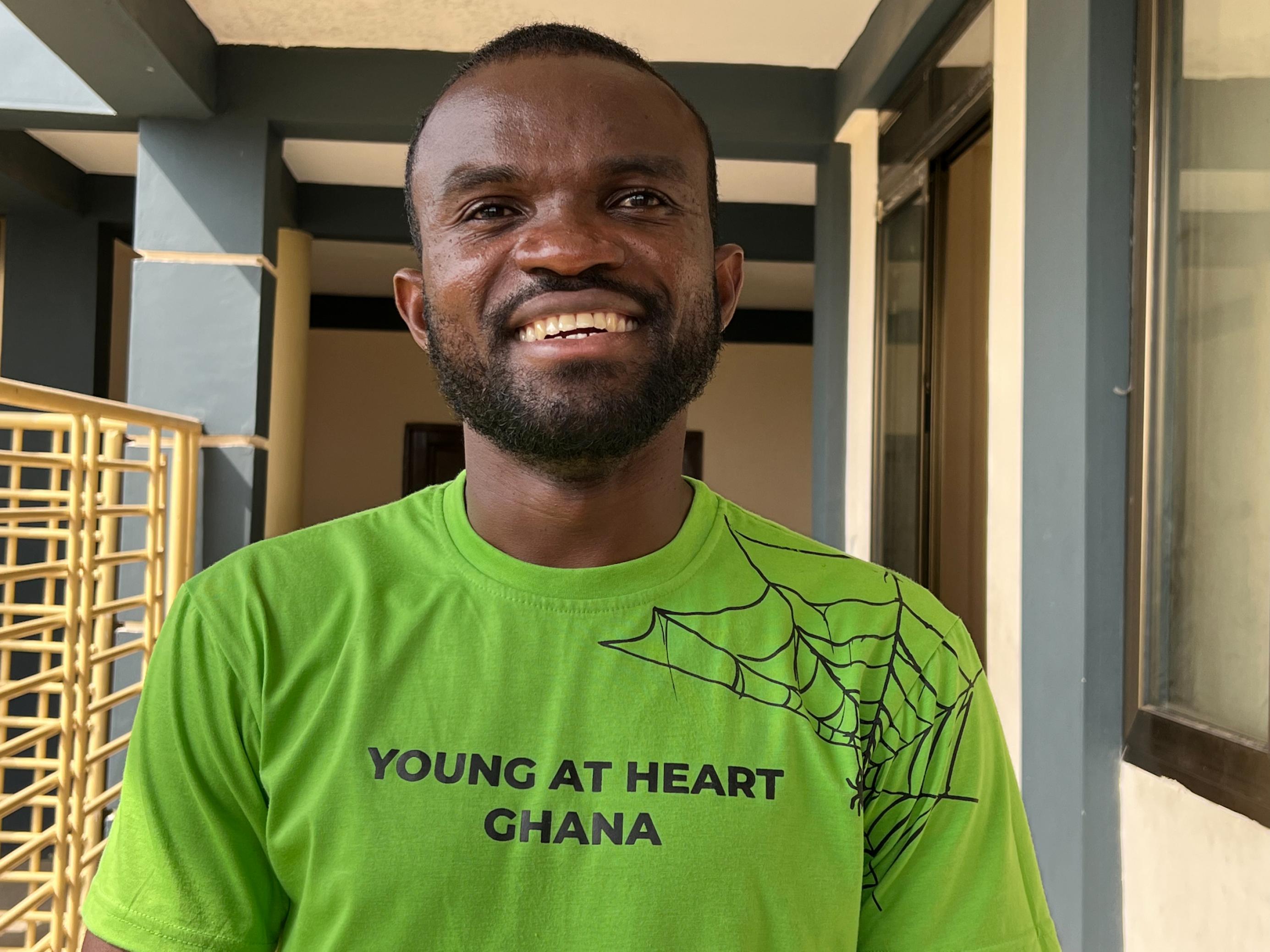 Ein junger Mann, breit lächelnd, trägt ein hellgrünes T-Shirt mit der Aufschrift Young At Heart Ghana.