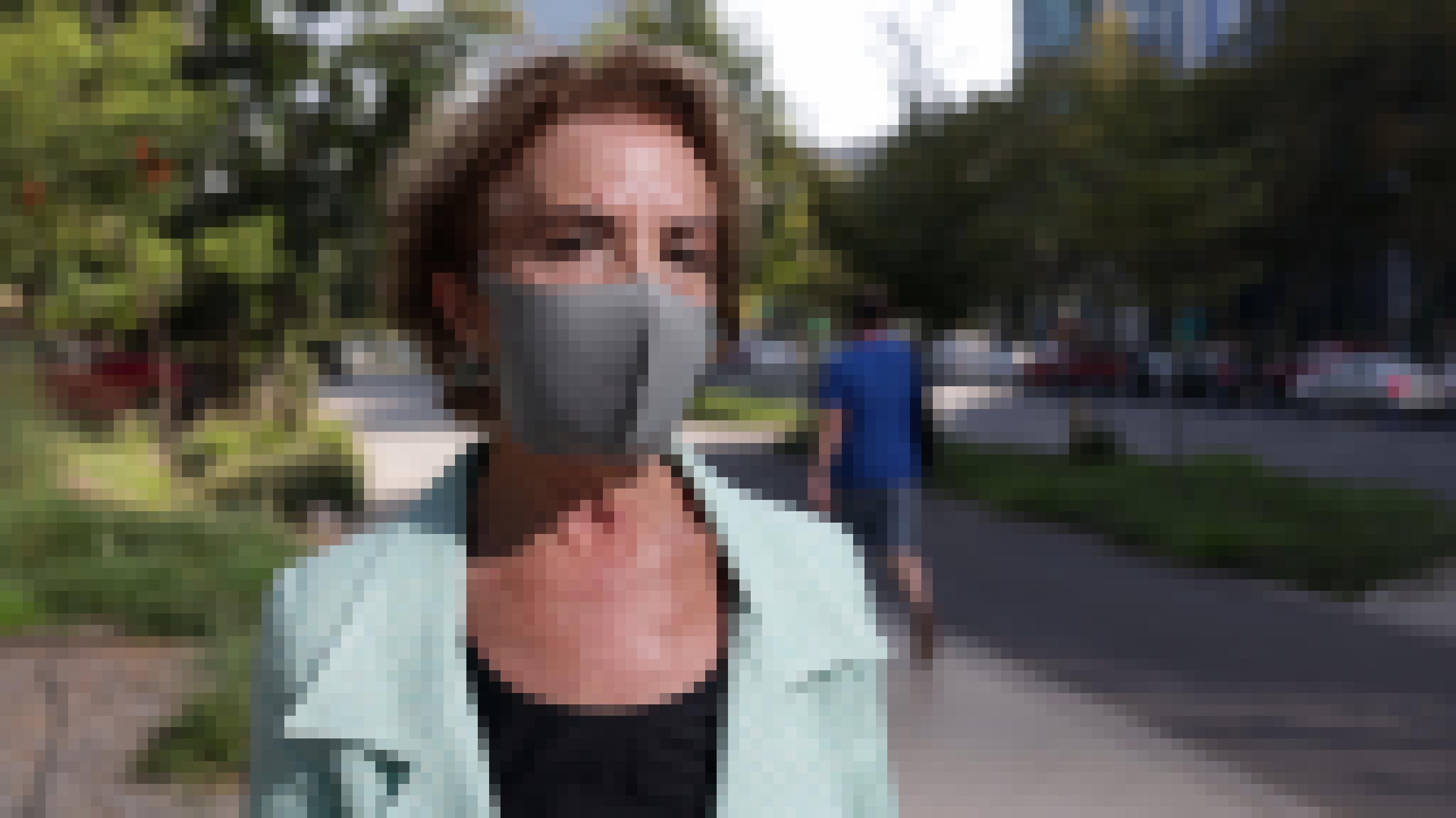 Eine Frau, die eine graue Gesichtsmaske trägt. Sie steht auf einem Gehweg in der Stadt.