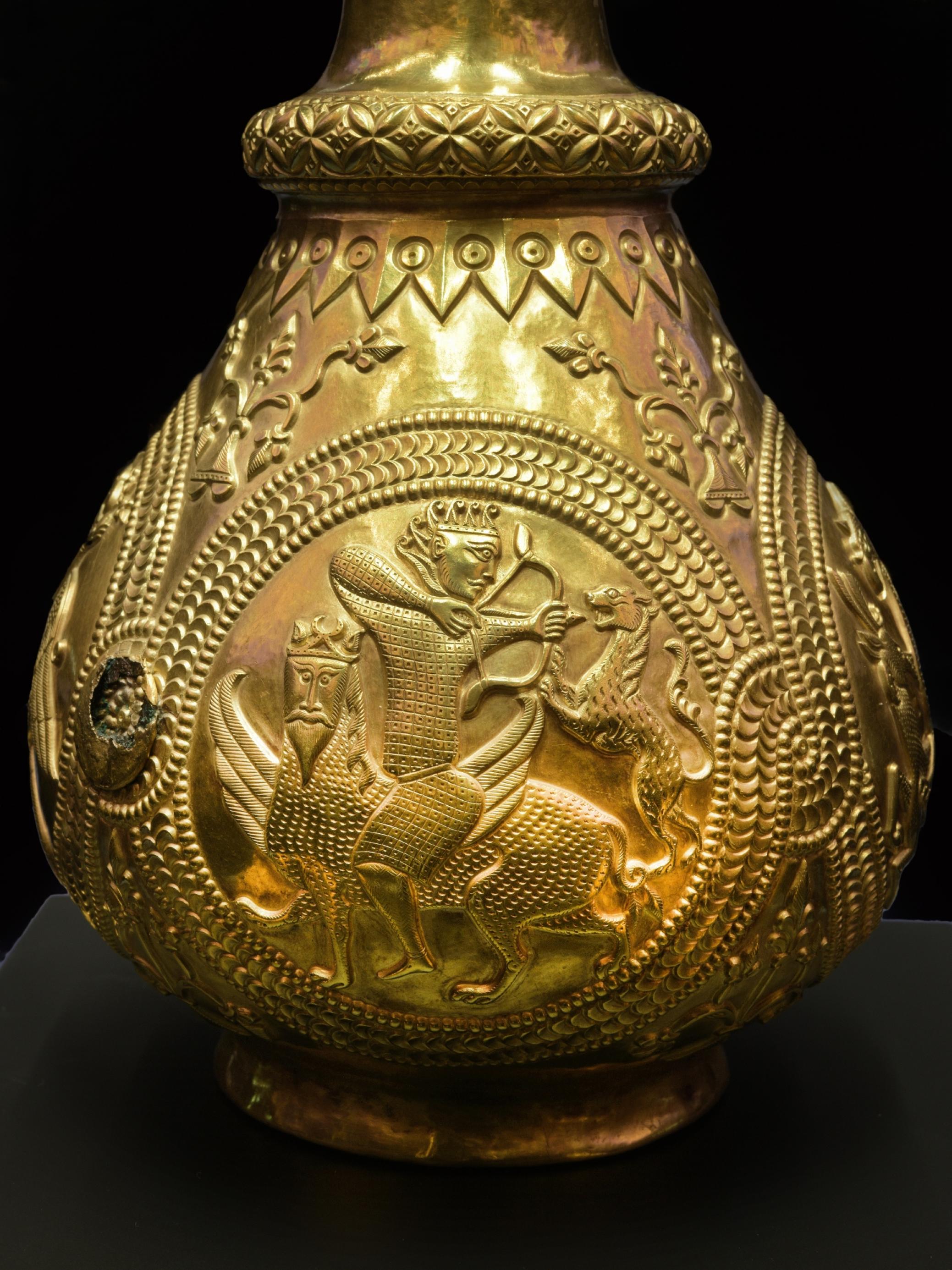 Das Bild zeigt eine goldene Kanne mit einem eingravierten Emblem, auf dem ein Mann ein Fabelwesen reitet und mit Pfeil und Bogen auf eine Raubkatze schießt.