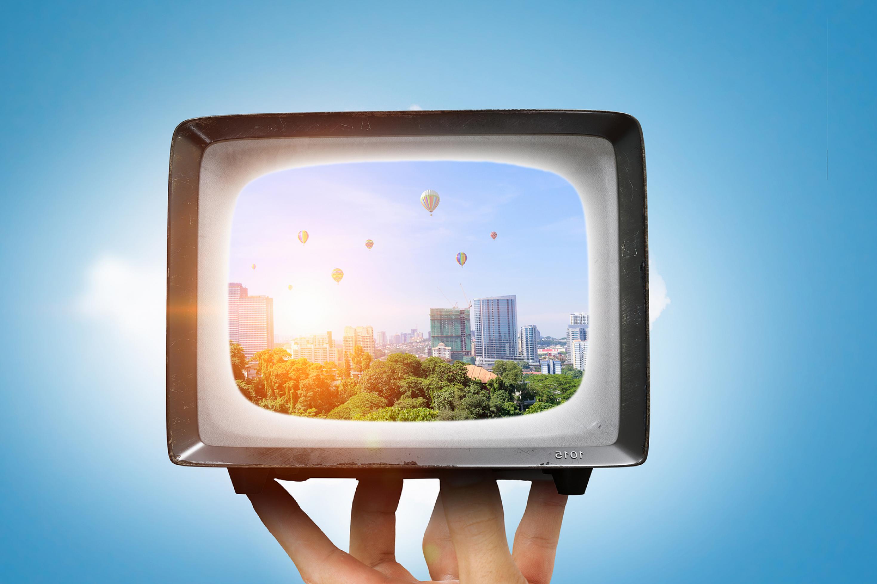 Symboldbild. Vor blauem Hintergrund hält eine Hand ein älteres Fernsehmodell in die Höhe. Darin zu sehen: eine grüne Stadt mit bunten Heißluftballons.