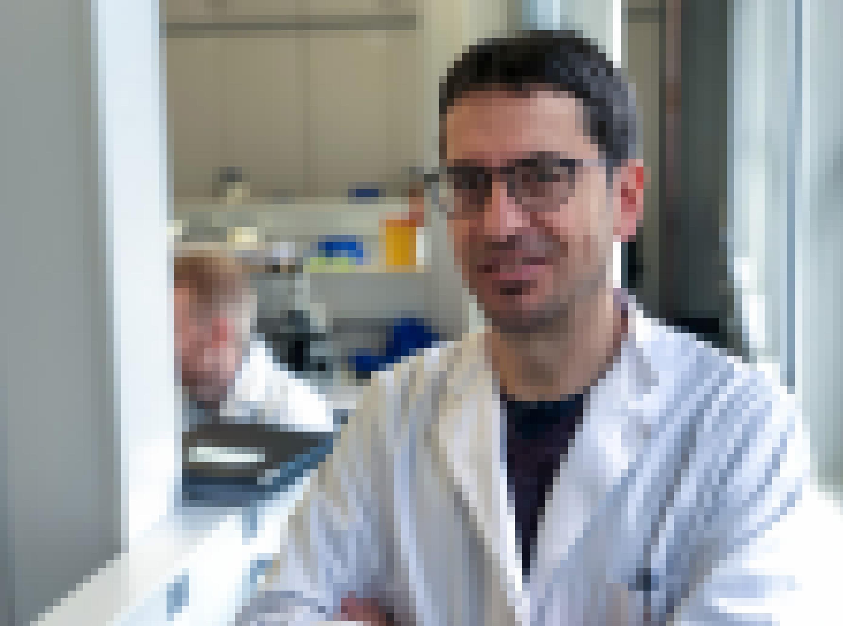 Ein dunkelhaariger Mann mit weißem Kittel und schwarzer Brille steht in einem Labor und schaut in die Kamera.