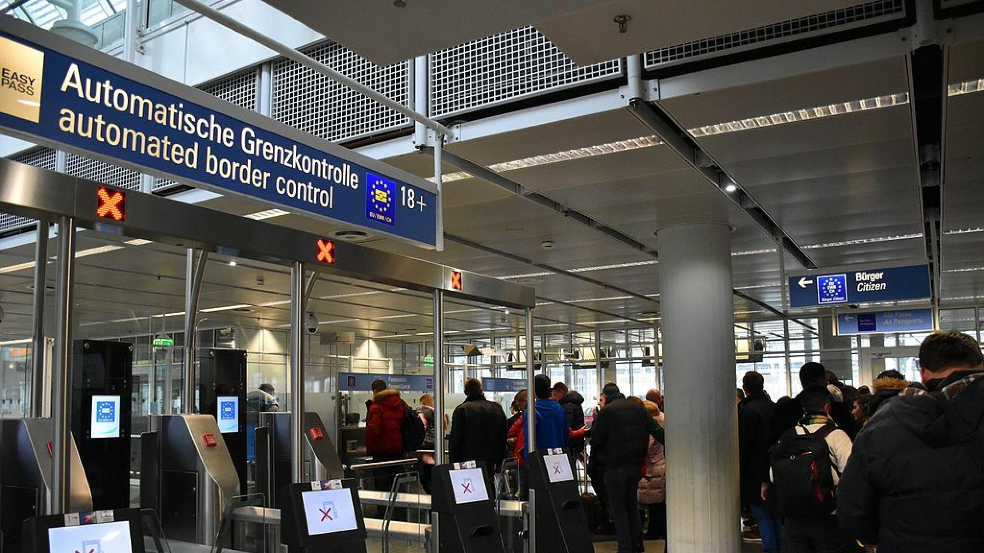 Menschen stehen am Flughafen an einer Passkontrolle in der Schlange.  Im Vordergrund sind leere Schalter für eine automatische Grenzkontrolle zu sehen.