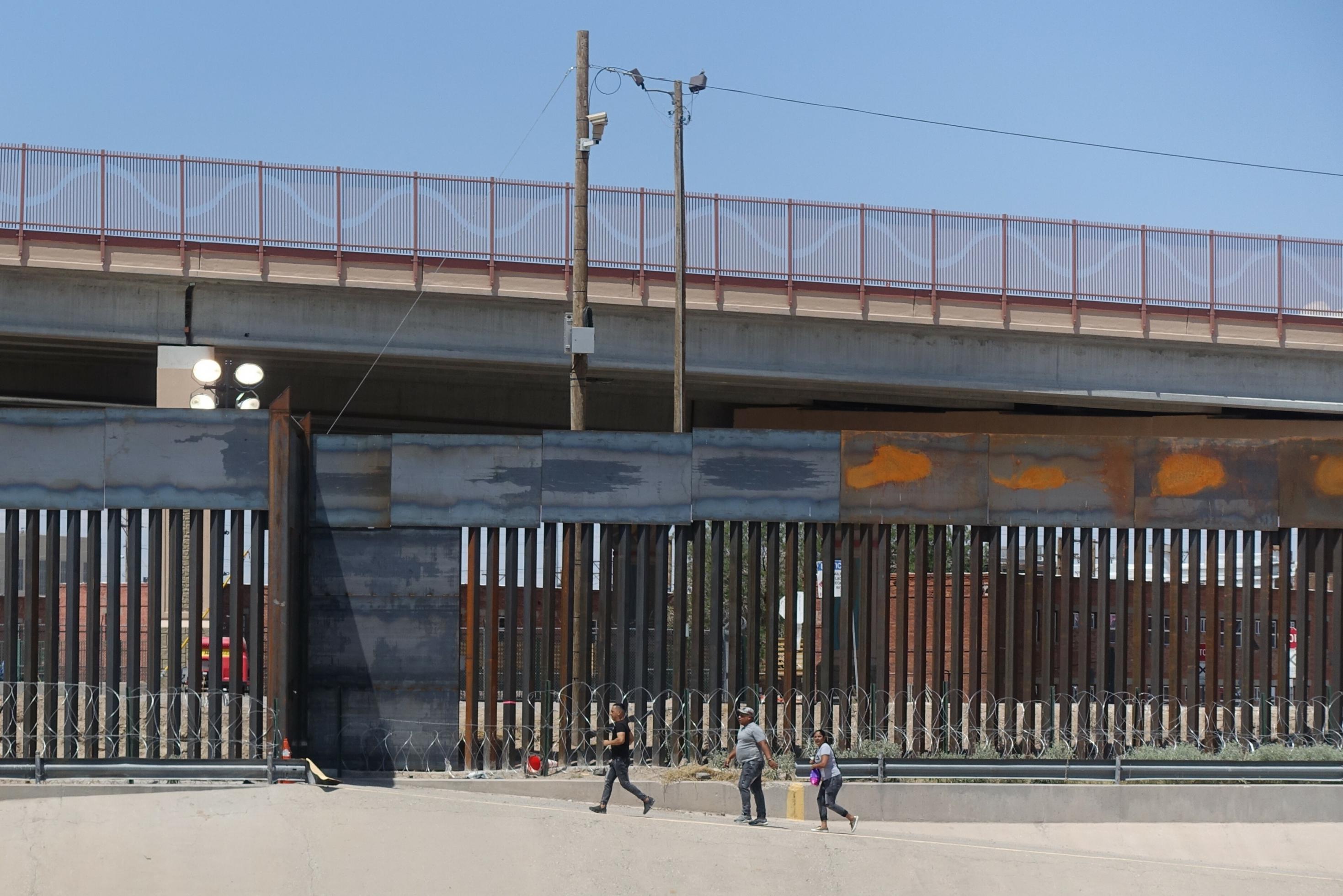 Das Foto zeigt die Grenzanlage im mexikanischen Ciudad Juarez, darüber ist eine Autobrücke zu sehen. Drei Passanten laufen an der Grenzmauer vorbei.