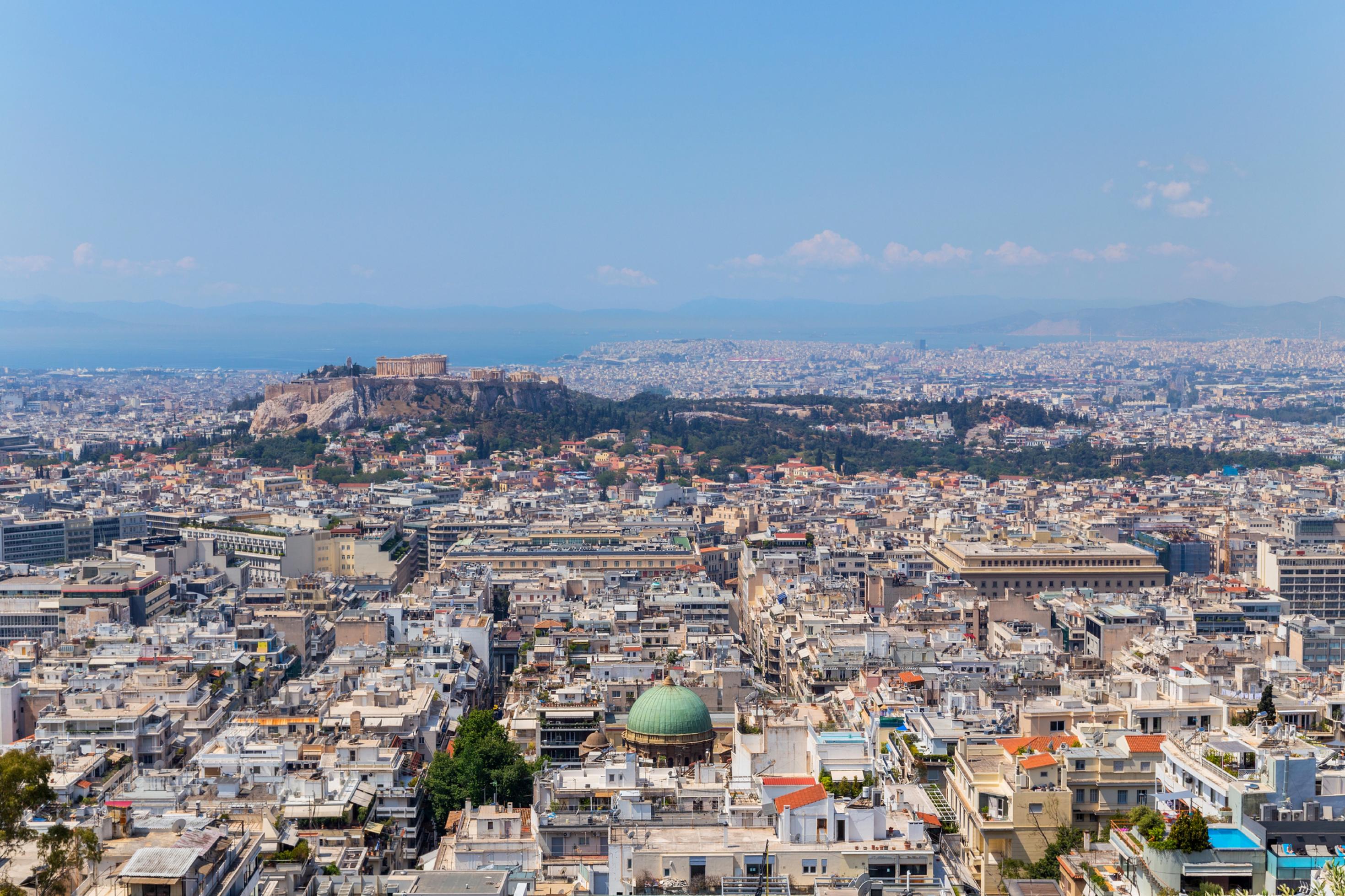 Das Stadtzentrum von Athen mit dicht gedrängten, hellen Gebäuden füllt das Bild. In der Mitte erhebt sich auf einem Hügel die Akropolis mit dem Parthenon, dahinter sieht man Meer, Berge und einen klaren blauen Himmel.