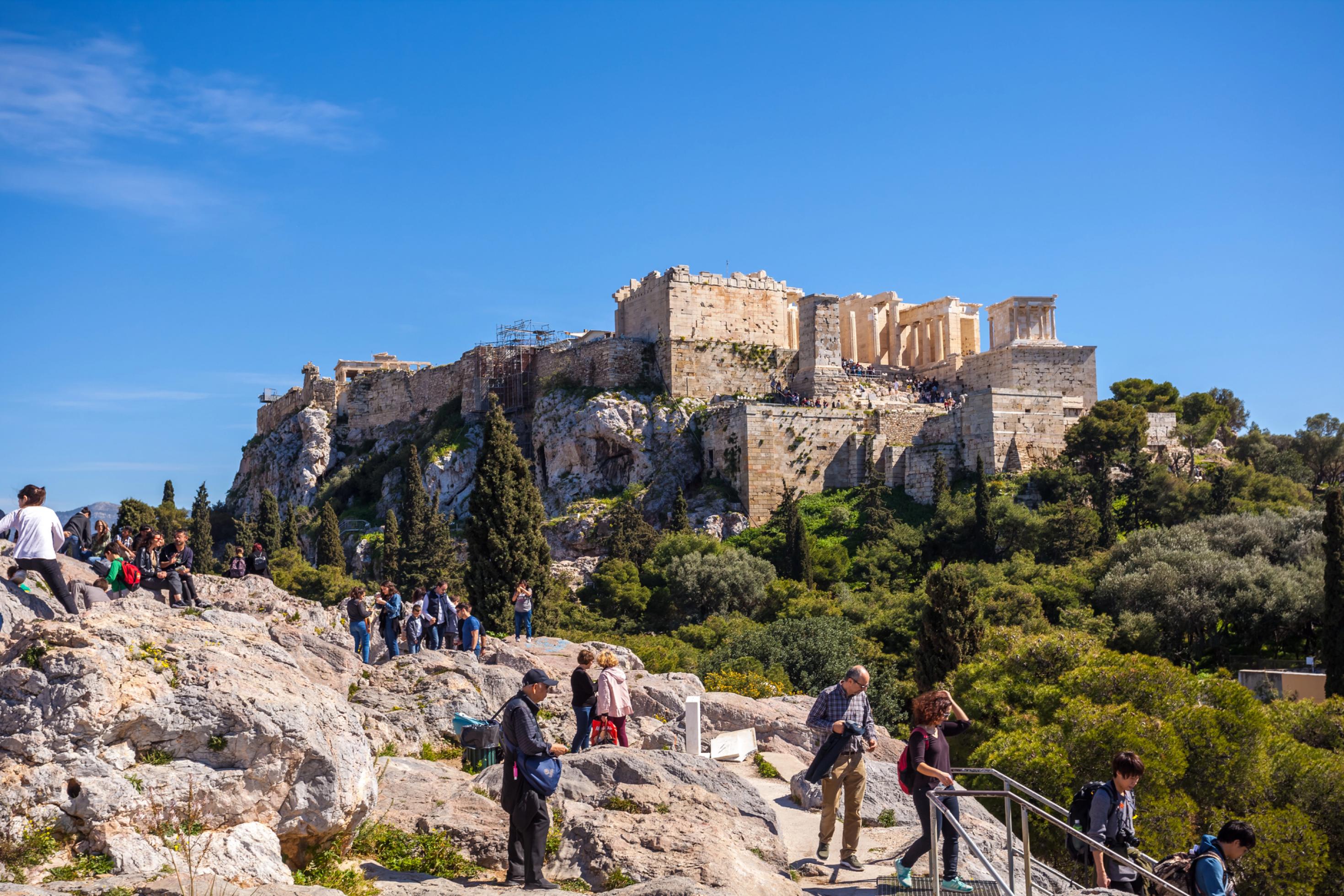 Zahlreiche Besucher halten sich auf einem Felsen auf, der vor der Akropolis von Athen liegt. Im Hintergrund ist die antike Festung auf einem höhergelegenen Felsen mit den bekannten Tempelruinen, darunter der Parthenon, zu sehen.
