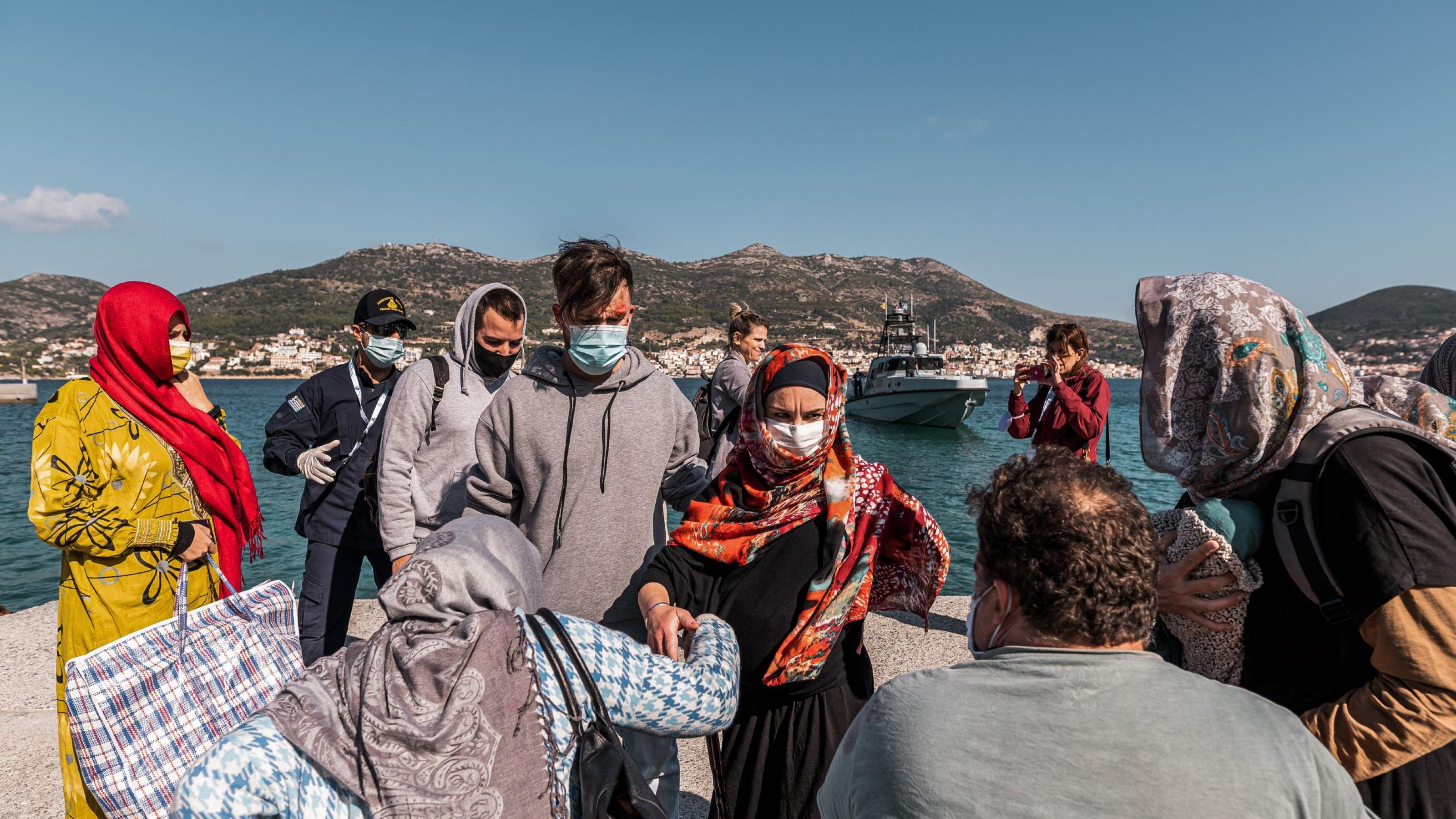 Das Bild zeigt eine Gruppe von Geflüchteten an einem sonnigen Hafen, begleitet von einem Küstenwache-Beamten. Viele tragen Masken, einige traditionelle Kleidung, und es ist ein Moment der Ankunft zu sehen. Im Hintergrund liegt eine Stadt vor einer bergigen Küstenlandschaft, während ein Patrouillenboot im Wasser liegt.