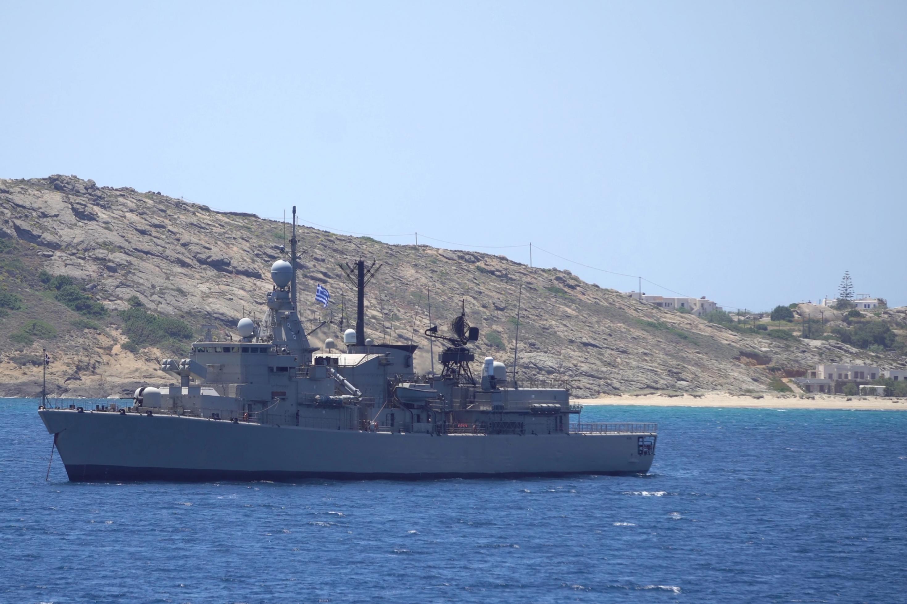 Ein Kriegsschiff der griechischen Marine fährt auf dem ruhigem, tiefblauen Meer. Im Hintergrund erhebt sich eine karge, felsige Inselküste mit wenigen hellen Gebäuden unter einem klaren, hellen Himmel.