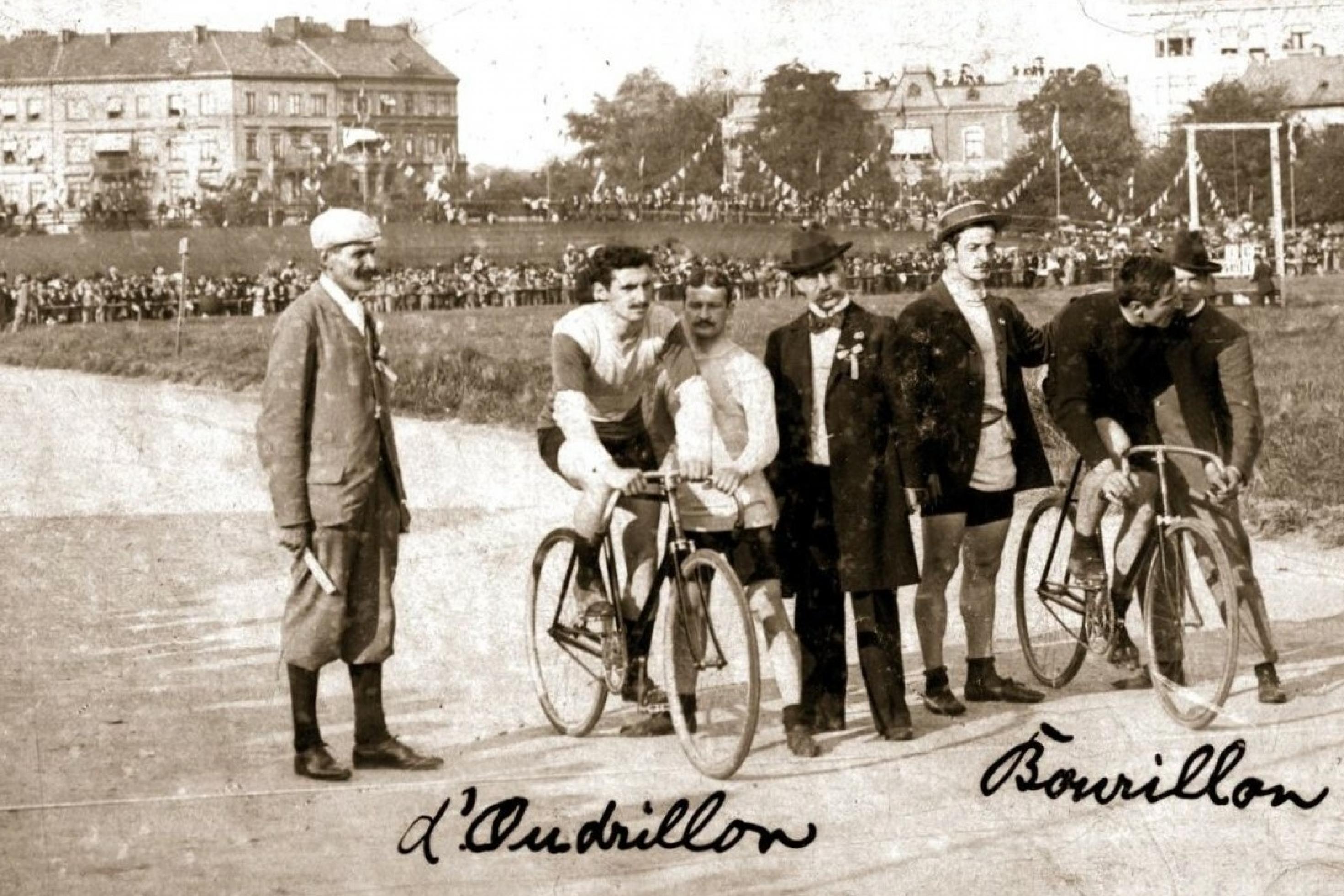 Das Schwarzweißfoto zeigt vor einem volksfestartigen Hintergrund zwei Rennfahrer auf ihren Fahrrädern am Start, Im Stand gehalten von Helfern.