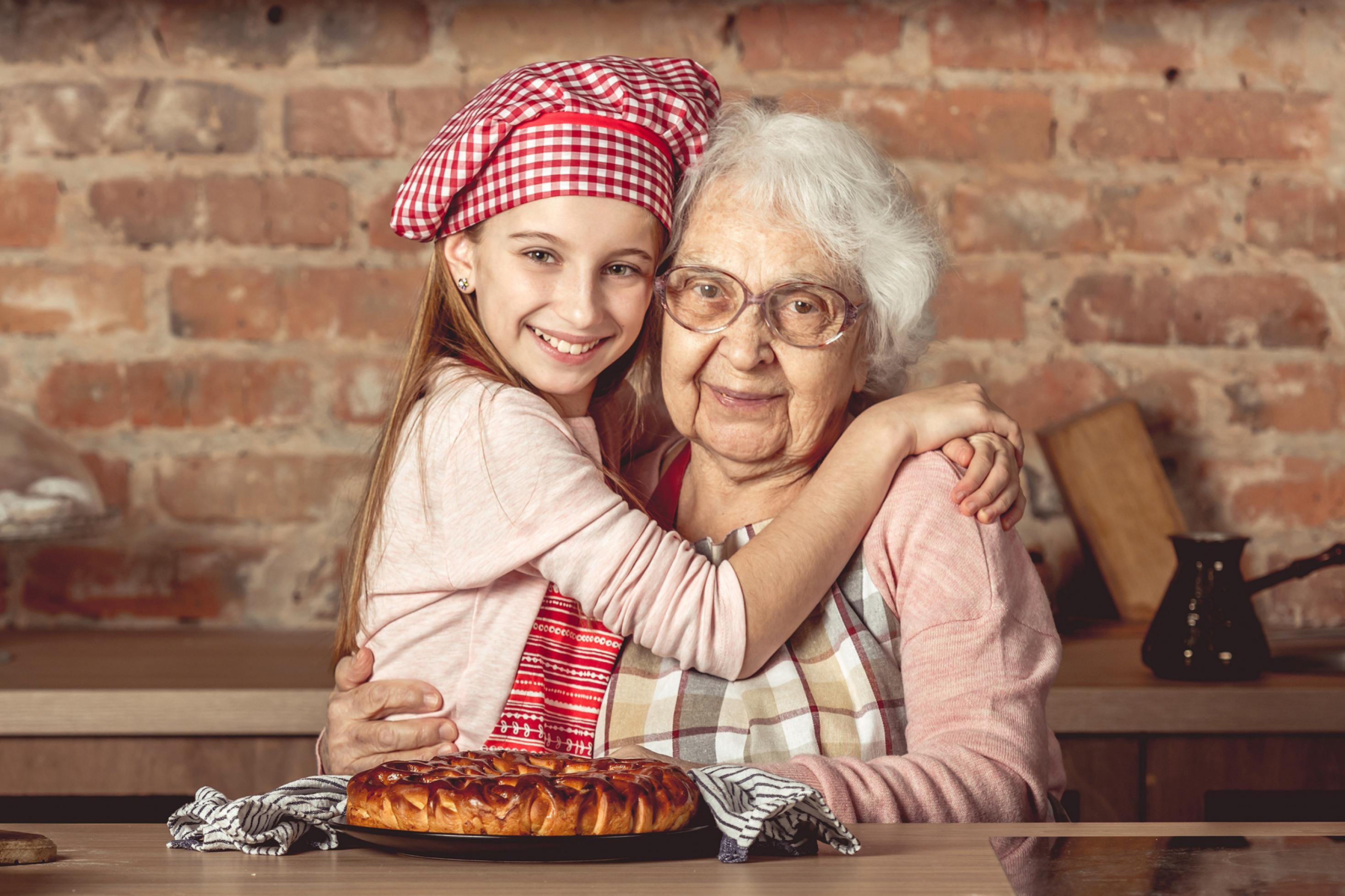 Das Foto zeigt eine weißhaarige, alte Frau mit Küchenschürze, die von einem lächelnden jungen Mädchen mit rot-weiß-karierter Kochmütze umarmt wird. Vor ihnen ein Gebäck. Sie befinden sich in einer altertümlichen Küche, hinter sich eine unverputzte Mauer aus roten Ziegelsteinen mit Ablage und Hängeschränken.