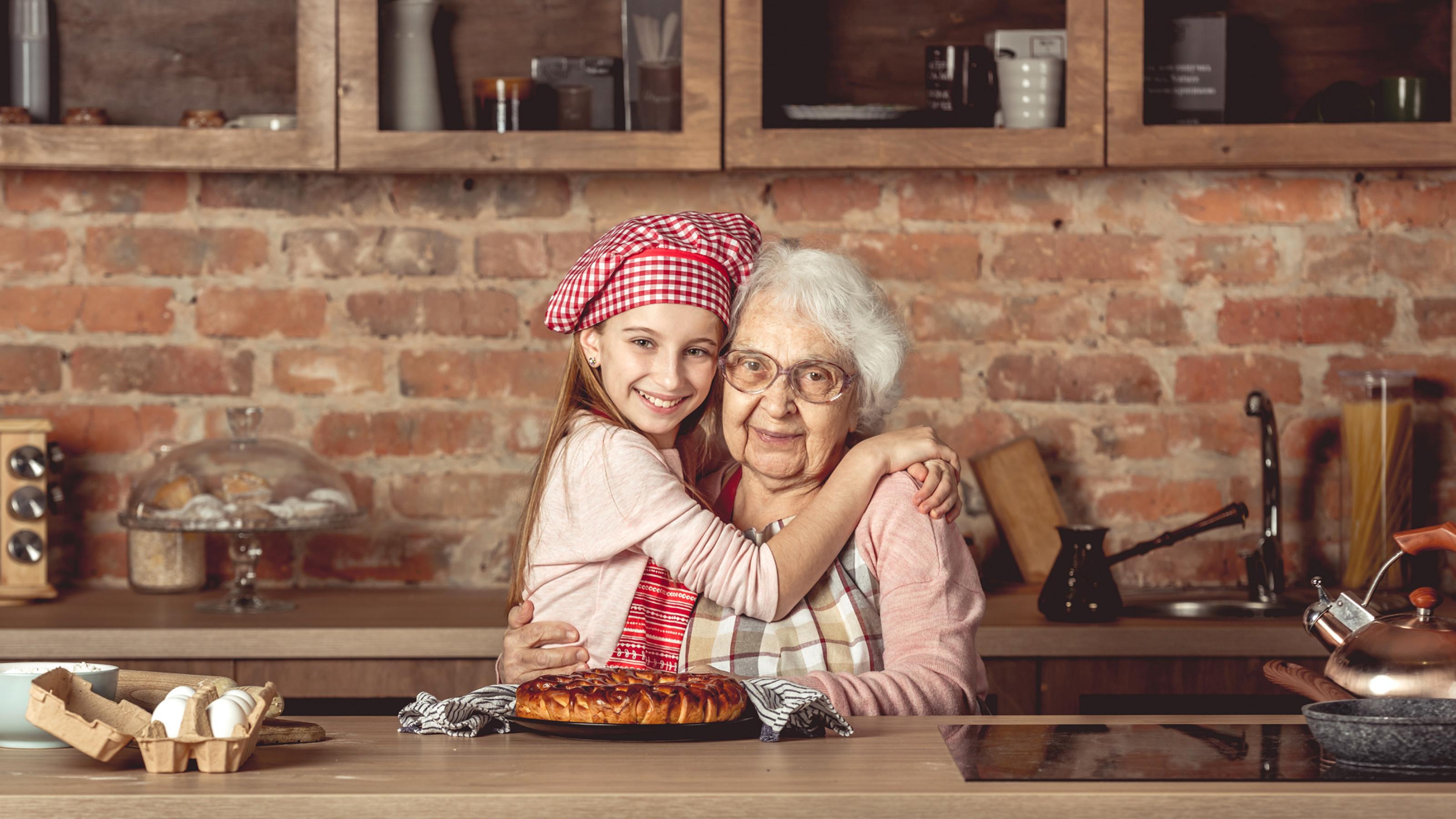 Das Foto zeigt eine weißhaarige, alte Frau mit Küchenschürze, die von einem lächelnden jungen Mädchen mit rot-weiß-karierter Kochmütze umarmt wird. Vor ihnen ein Gebäck. Sie befinden sich in einer altertümlichen Küche, hinter sich eine unverputzte Mauer aus roten Ziegelsteinen mit Ablage und Hängeschränken.
