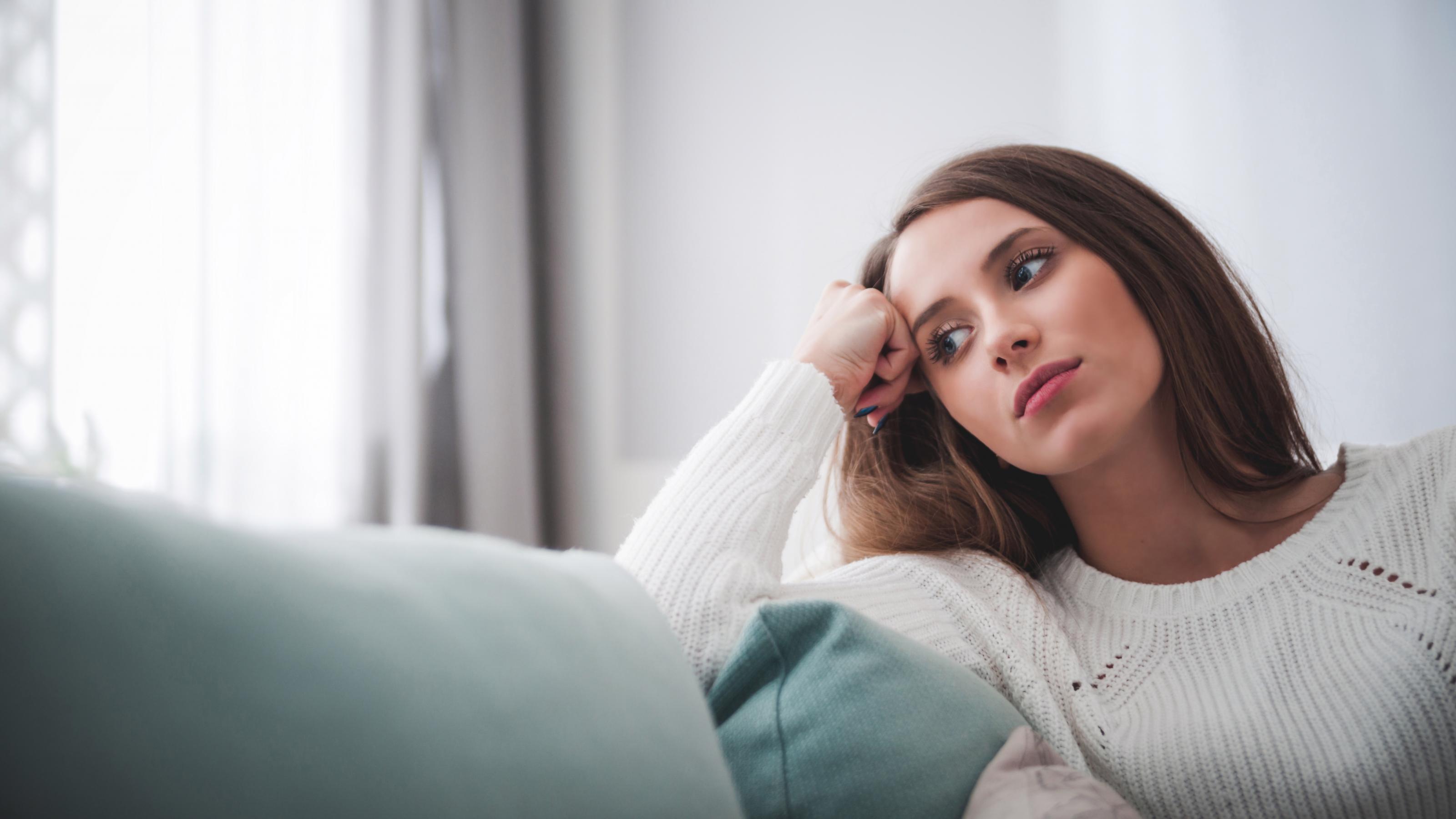 Eine junge Frau sitzt auf einem grauen Sofa, stützt den Kopf auf einem Arm ab und blickt ins Nirgendwo. Sie sieht nachdenklichh aus.