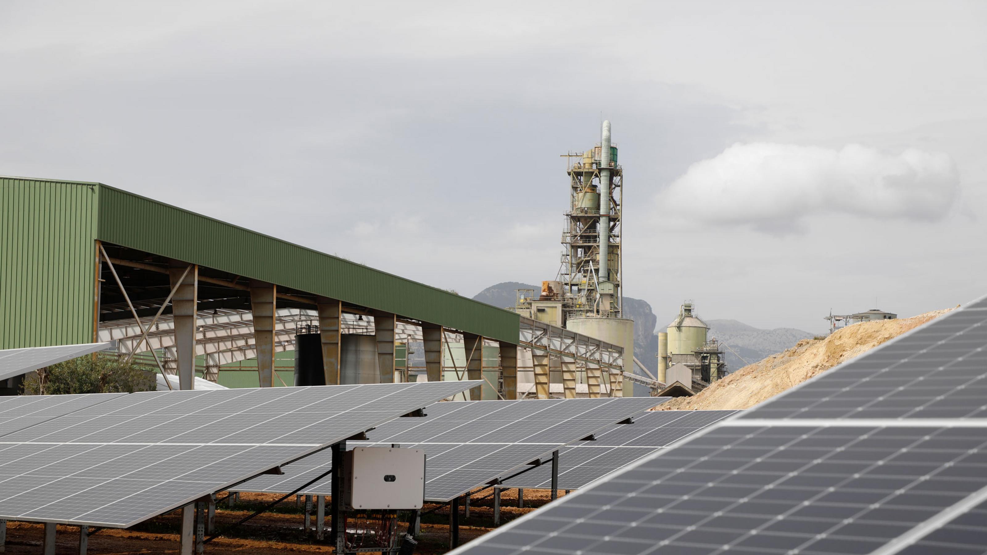 Auf dem Bild ist eine Photovoltaikanlage zu sehen, im Hintergrund stehen Fabrikschlote. Mit Hilfe von Solarstrom wird auf Mallorca in einer ehemaligen Zementfabrik grüner Wasserstoff produziert.