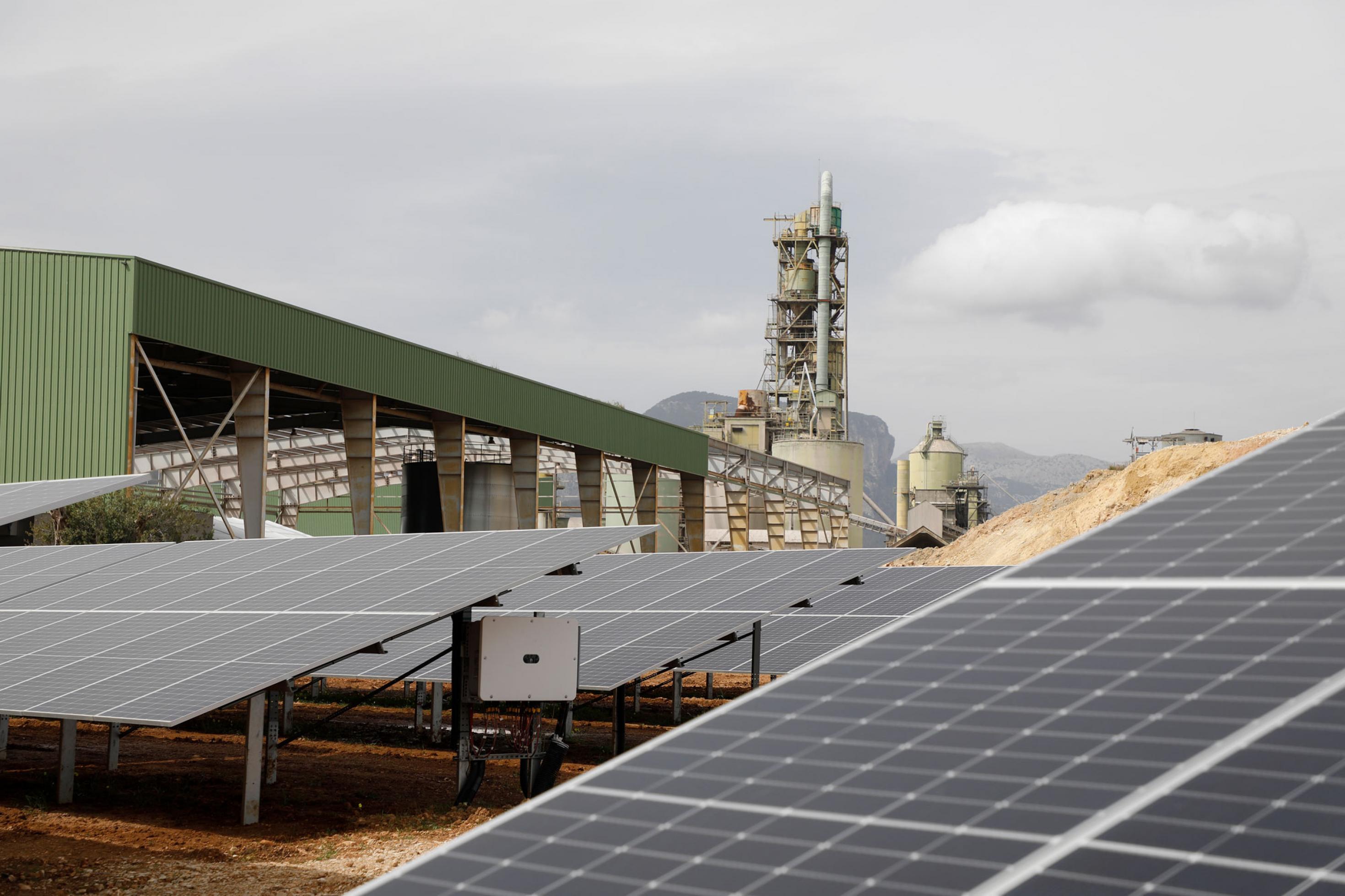 Auf dem Bild ist eine Photovoltaikanlage zu sehen, im Hintergrund stehen Fabrikschlote. Mit Hilfe von Solarstrom wird auf Mallorca in einer ehemaligen Zementfabrik grüner Wasserstoff produziert.