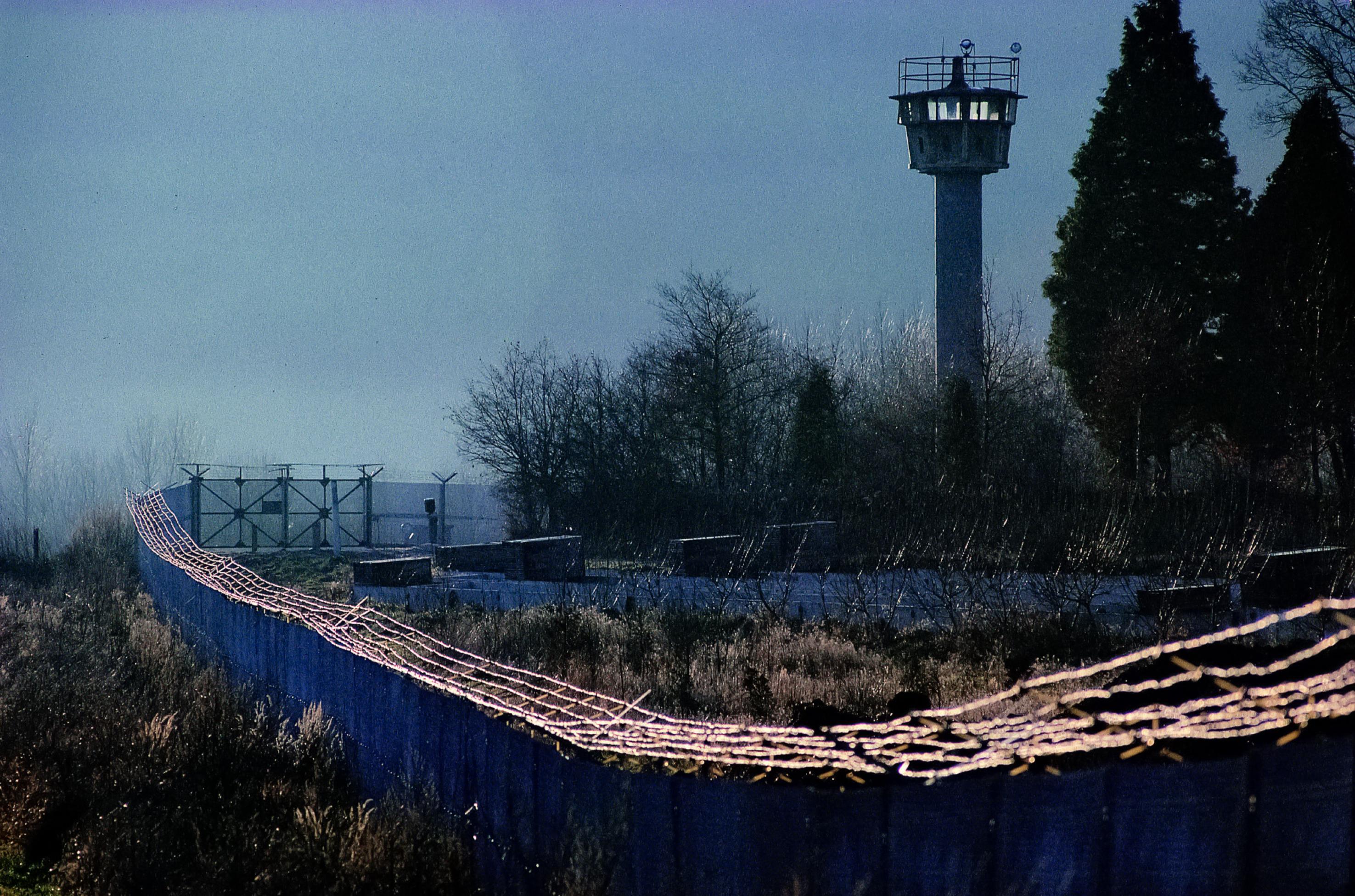 Stacheldrahtbewehrte Zäune ziehen sich vor einem DDR-Wachturm entlang. Die Anlage wirkt brutal.