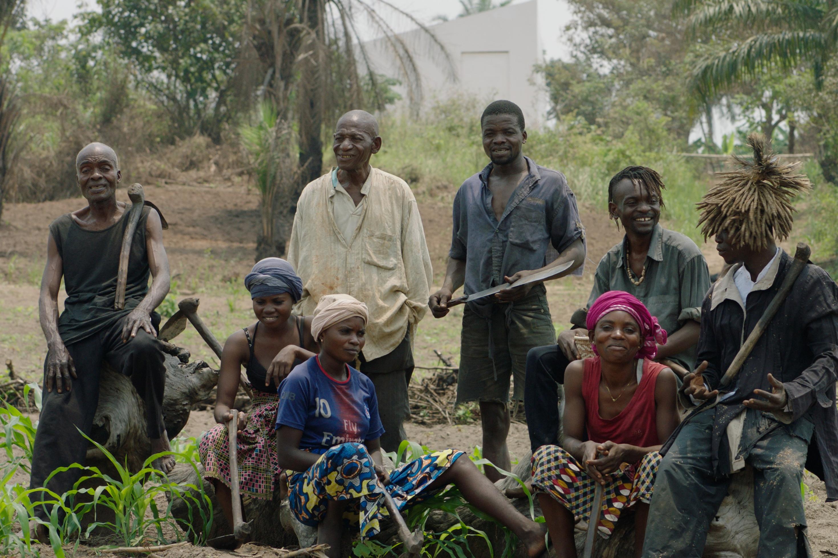 Kongolesische Männer und Frauen posieren mit Hacken vor der Kulisse eines schütter bewachsenen Landstrichs. Im Hintergrund ist ein modernes weißes Gebäude zu sehen.