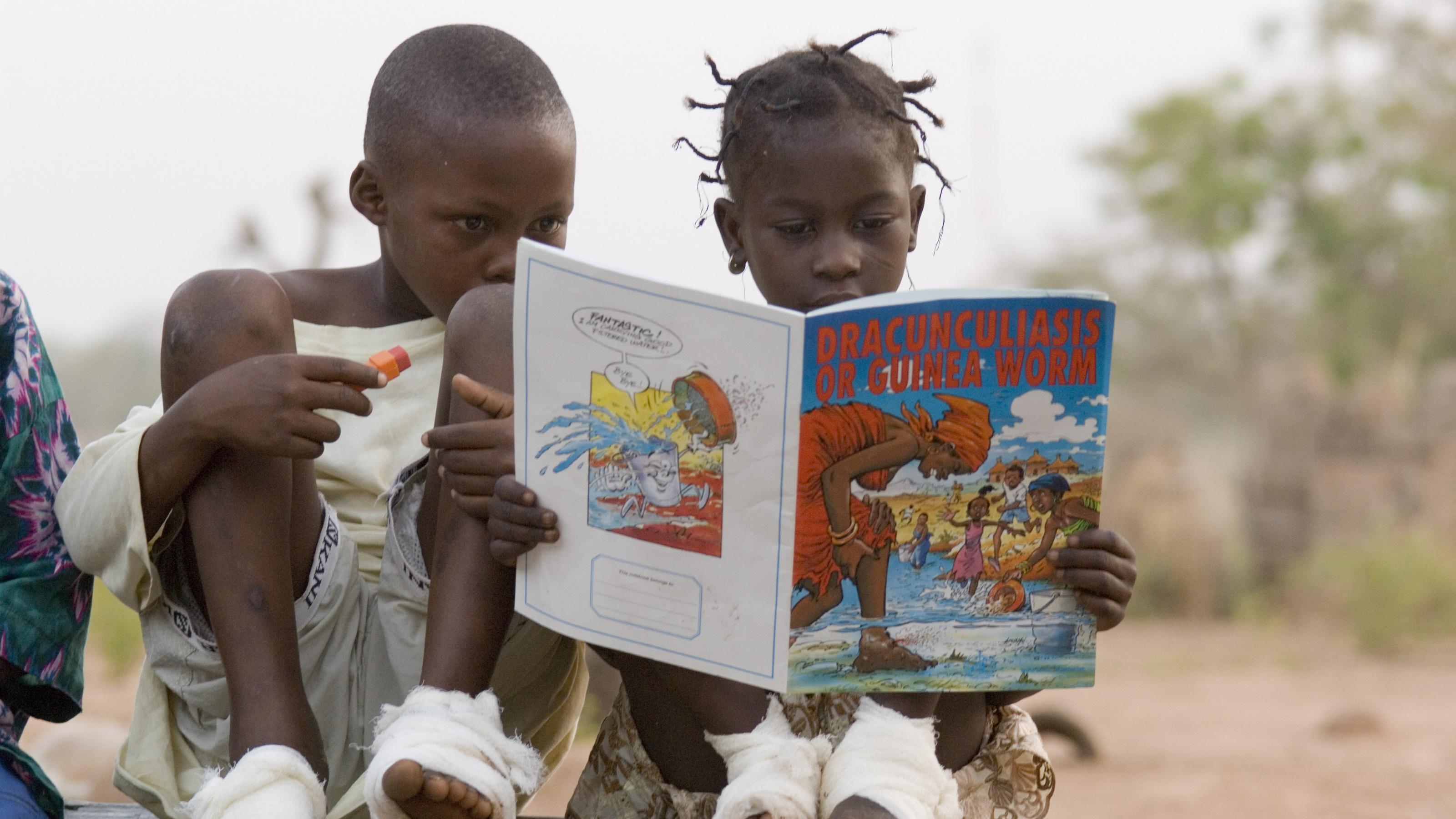 Zwei Kinder mit bandagierten Füßen lesen in einem Heft, in dem es um den Guineawurm geht.