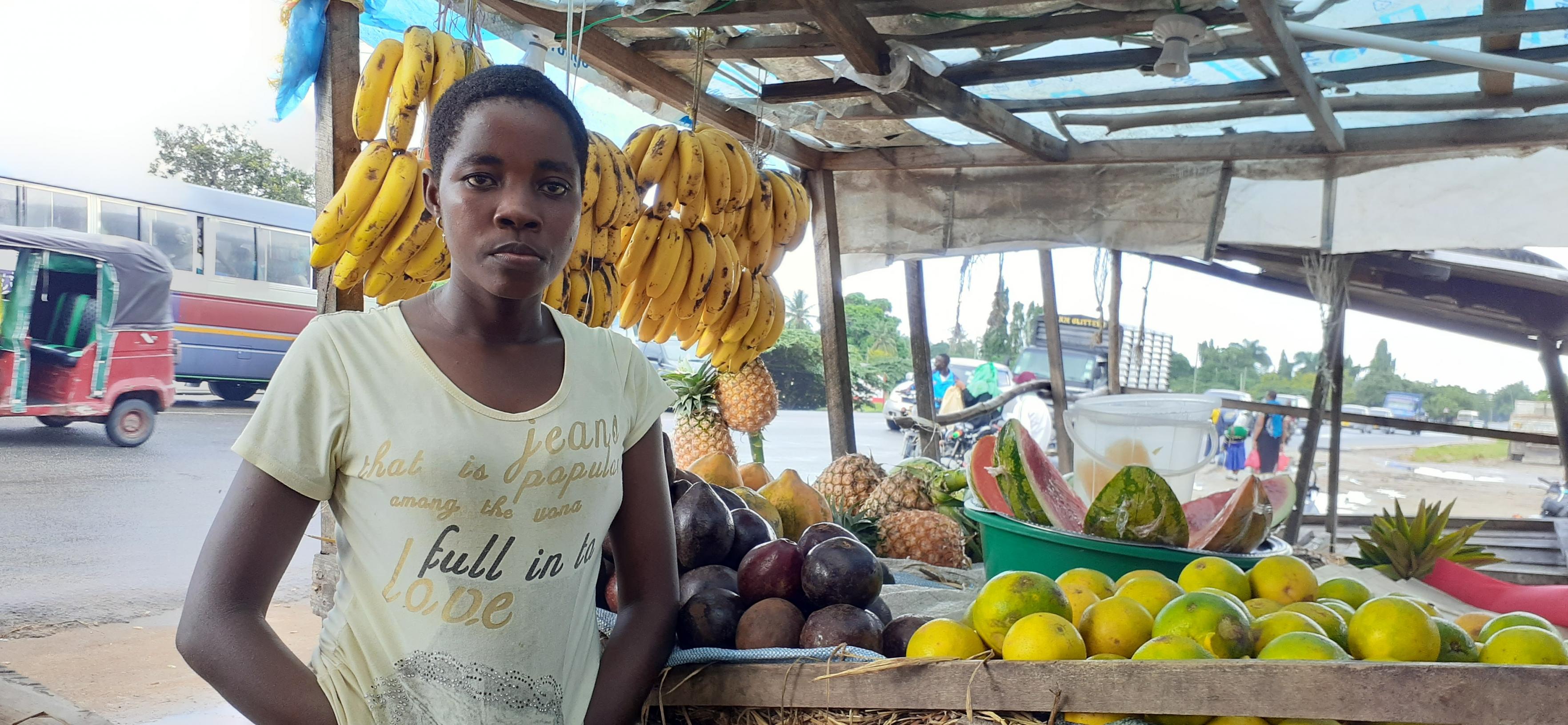Eine junge Frau steht vor ihrem Marktstand. Bananen, Ananas, Papaya und Avocados sind sorgfältig angeordnet.