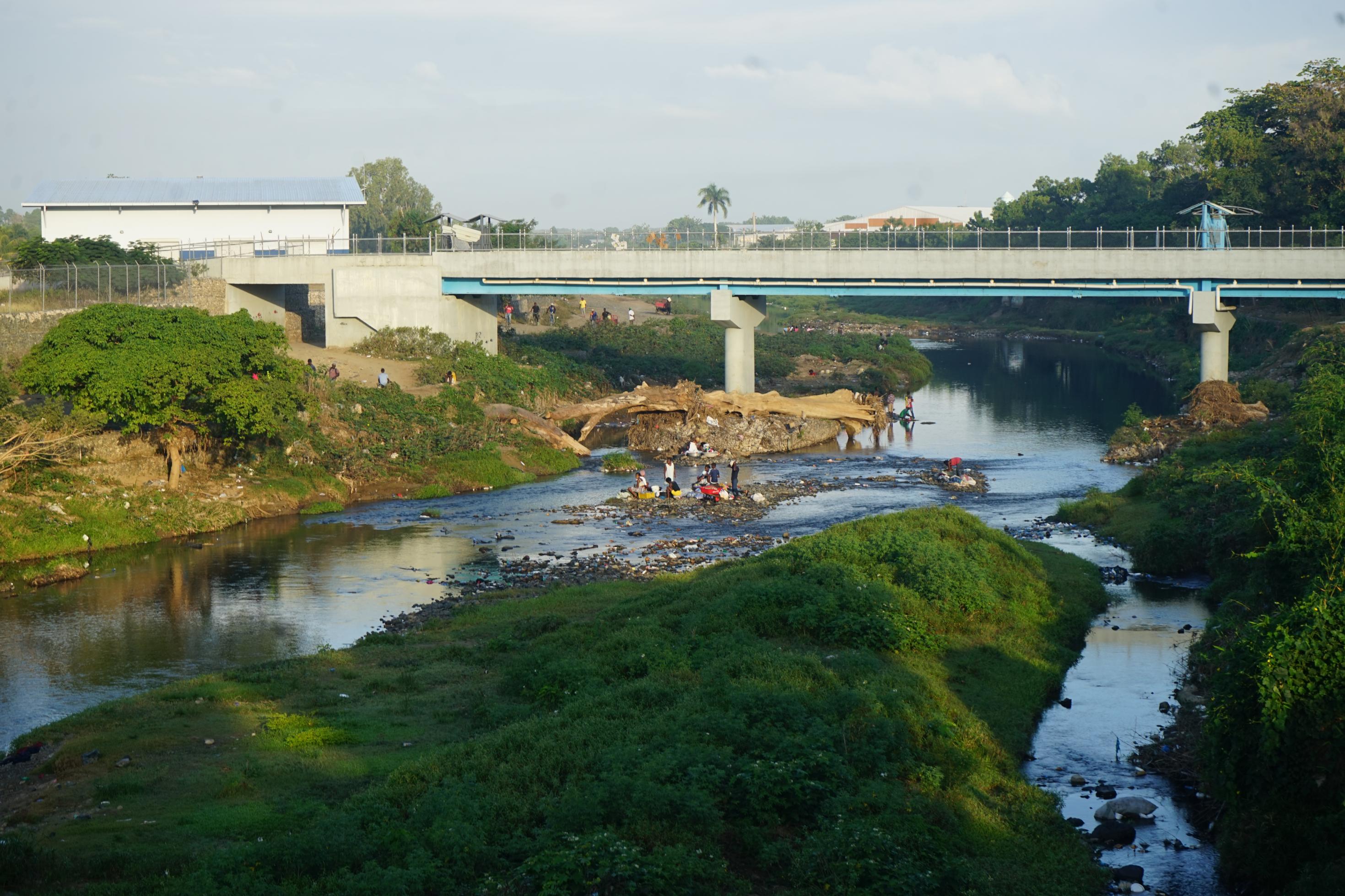 Ein Fluss, in dem Frauen Wäsche waschen, im Hintergrund eine BrÜcke.