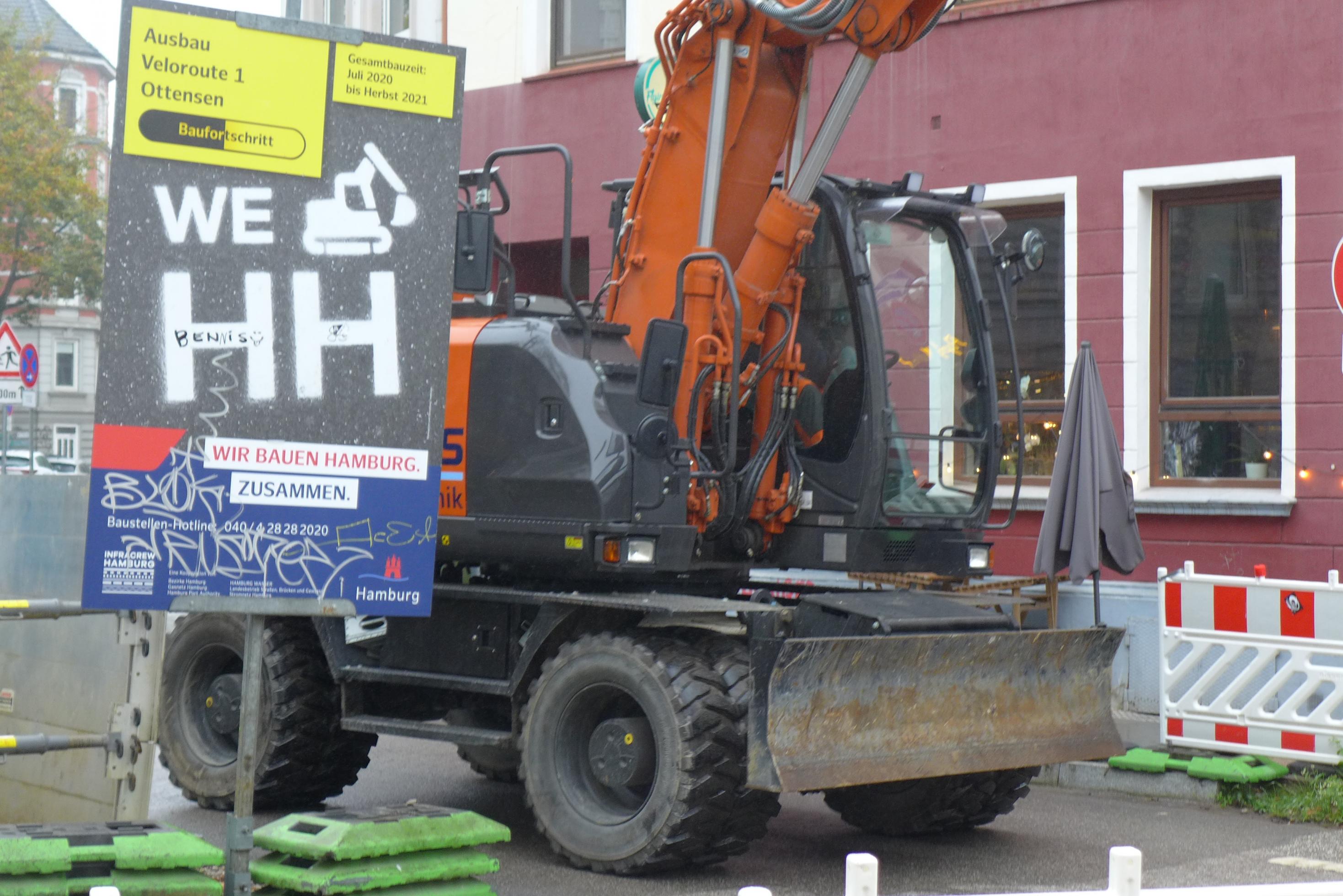 Ein orangefarbener mittelgroßer Schaufelbagger fährt hinter einem Schild vorbei. Die Tafel beschreibt den Baufortschritt der Veloroute Hamburg. Auf einem gelben Papier steht: Ausbau Veloroute 1 Ottensen. Unten steht in rot und blau: Wir bauen Hamburg – zusammen. Das Schild ist mit Graffiti beschmiert.