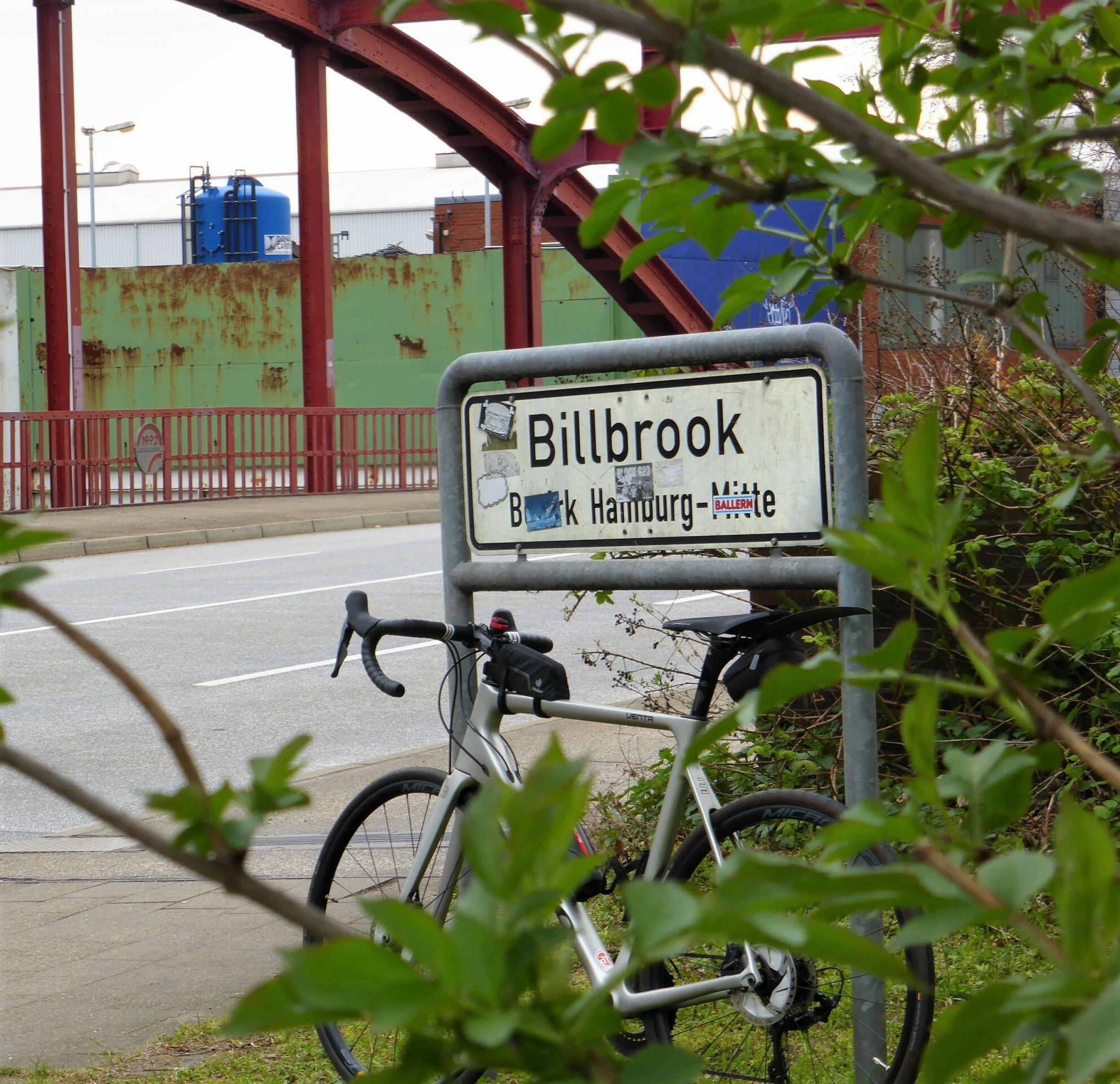 Im Hintergrund zeichnen sich rostrote Eisenbögen einer Brücke ab. Im Vordergrund zu sehen ist das Schild, das den Beginn des Stadtteils Billbrook markiert. Daran lehnt das Rennrad des RadelndenReporters.