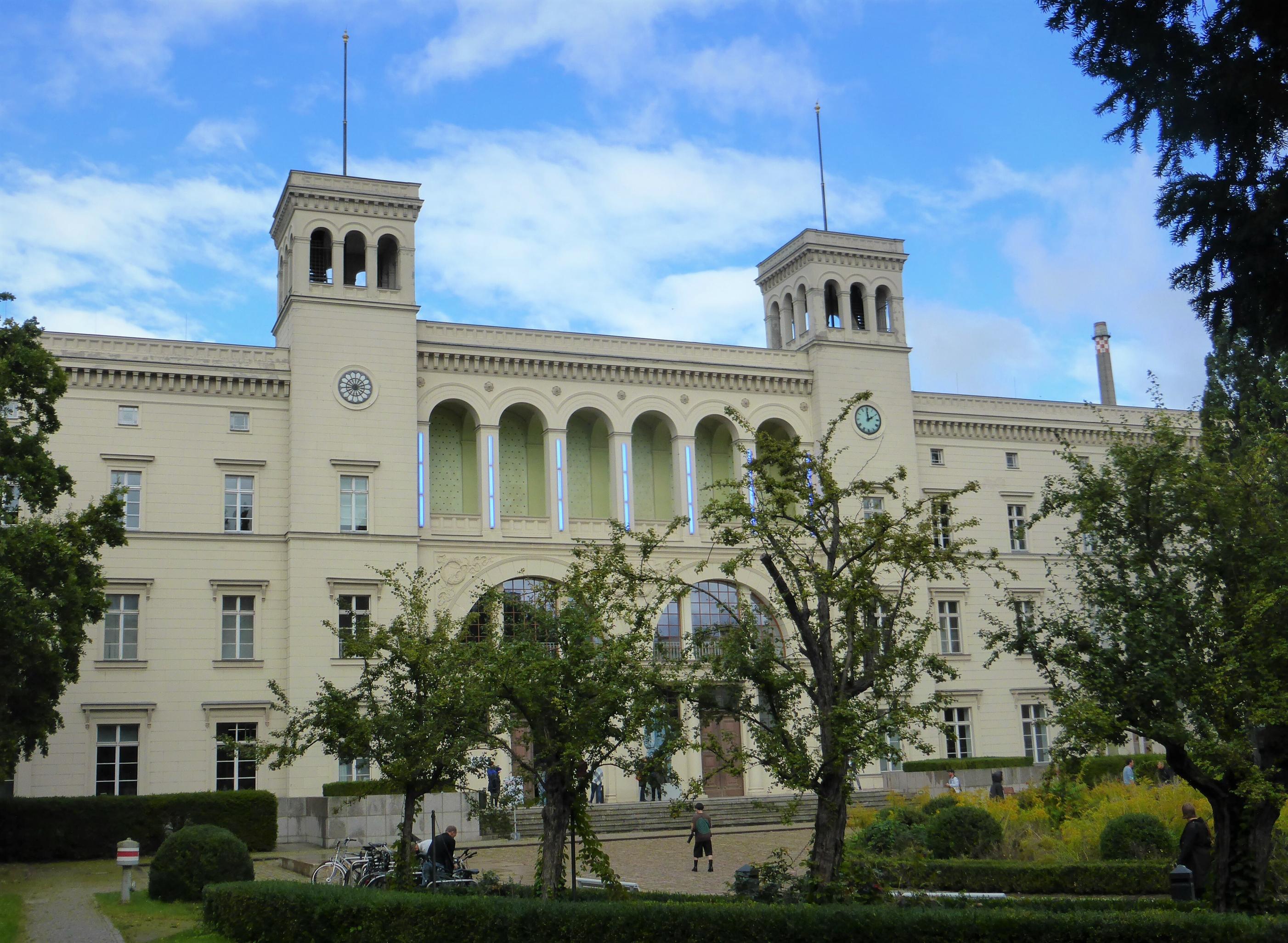 Hinter Bäumen und Büschen ragt die von zwei Turmbauten bekrönte Schaufassade des klassizistischen Bahnhofsgebäudes auf, deren Mittelteil wie ein Stadttor wirkt.