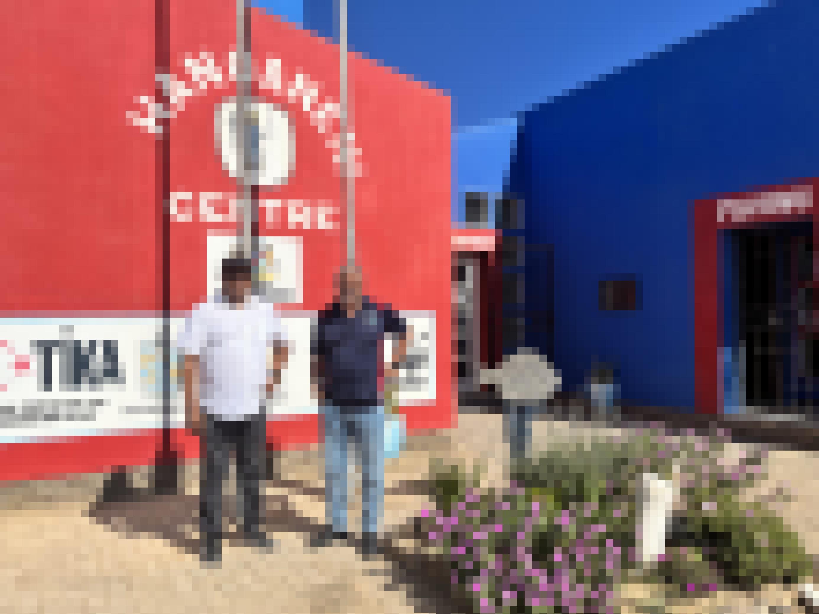 Die beiden Männer stehen vor dem rot-blauen HAFA-Gebäude. Eine Hand in die Hüfte gestemmt.