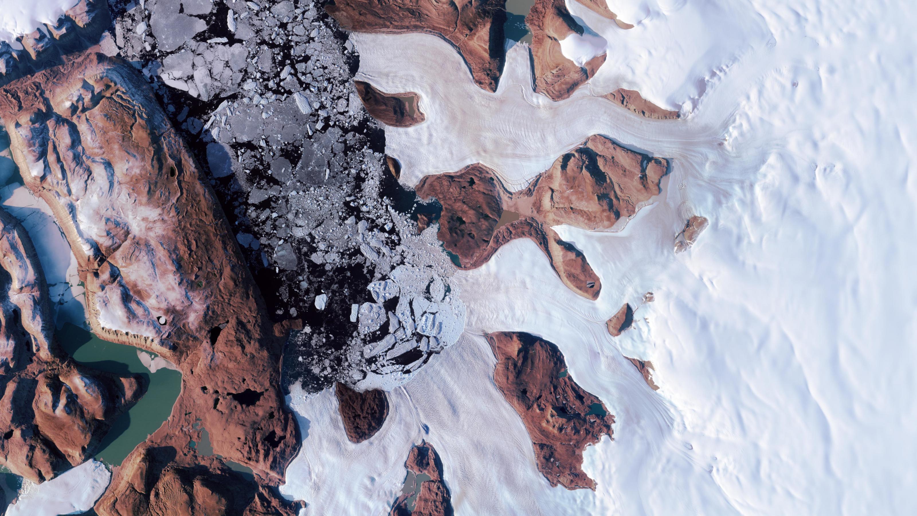 Satellitenbild einer arktischen Landschaft: braune Felsen in einer weißen eisigen Landschaft, links ein länglicher Wasserkörper mit zerbrochenen Eisbergen in dunkelblauem Wasser. Das Foto wirkt künstlerisch.