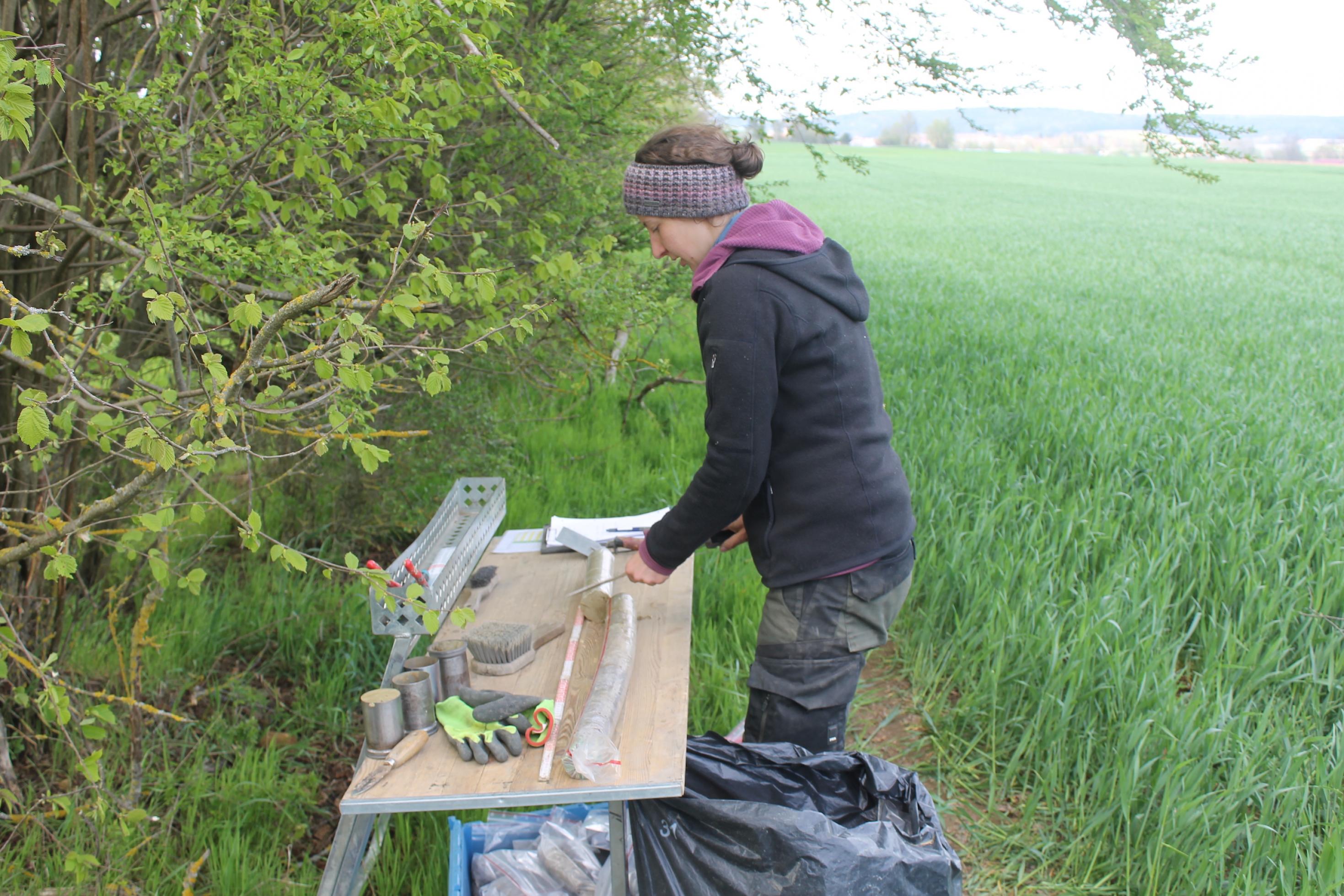 Eine junge Frau in lila Jacke und mit Stirnband steht an einem Tapeziertisch an einem Waldrand. Auf dem Tisch befinden sich Forschungsutensilien.