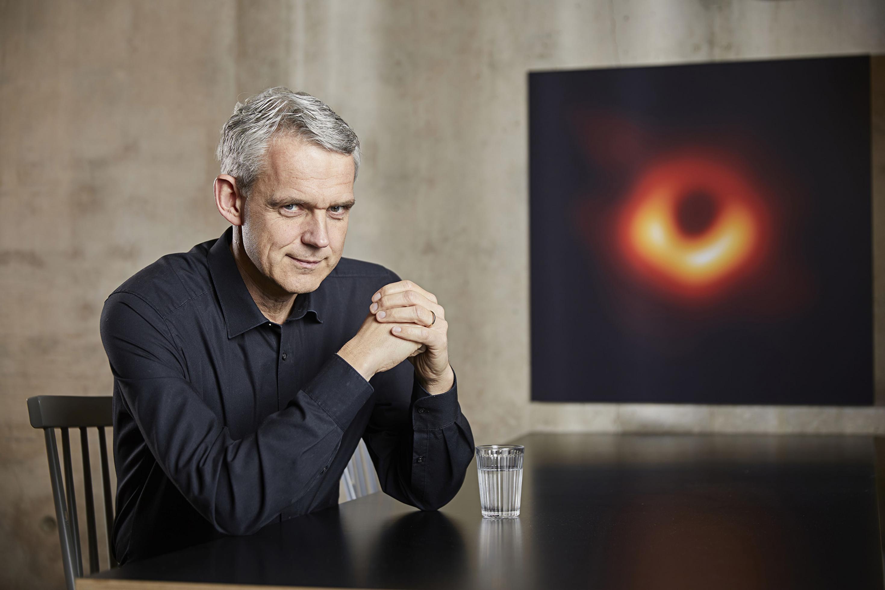 Heino Falcke am Tisch sitzend. Im Hintergrund hängt ein Bild des Schwarzen Lochs M87*, das er und seine Kollegen und Kolleginnen aufgenommen haben.