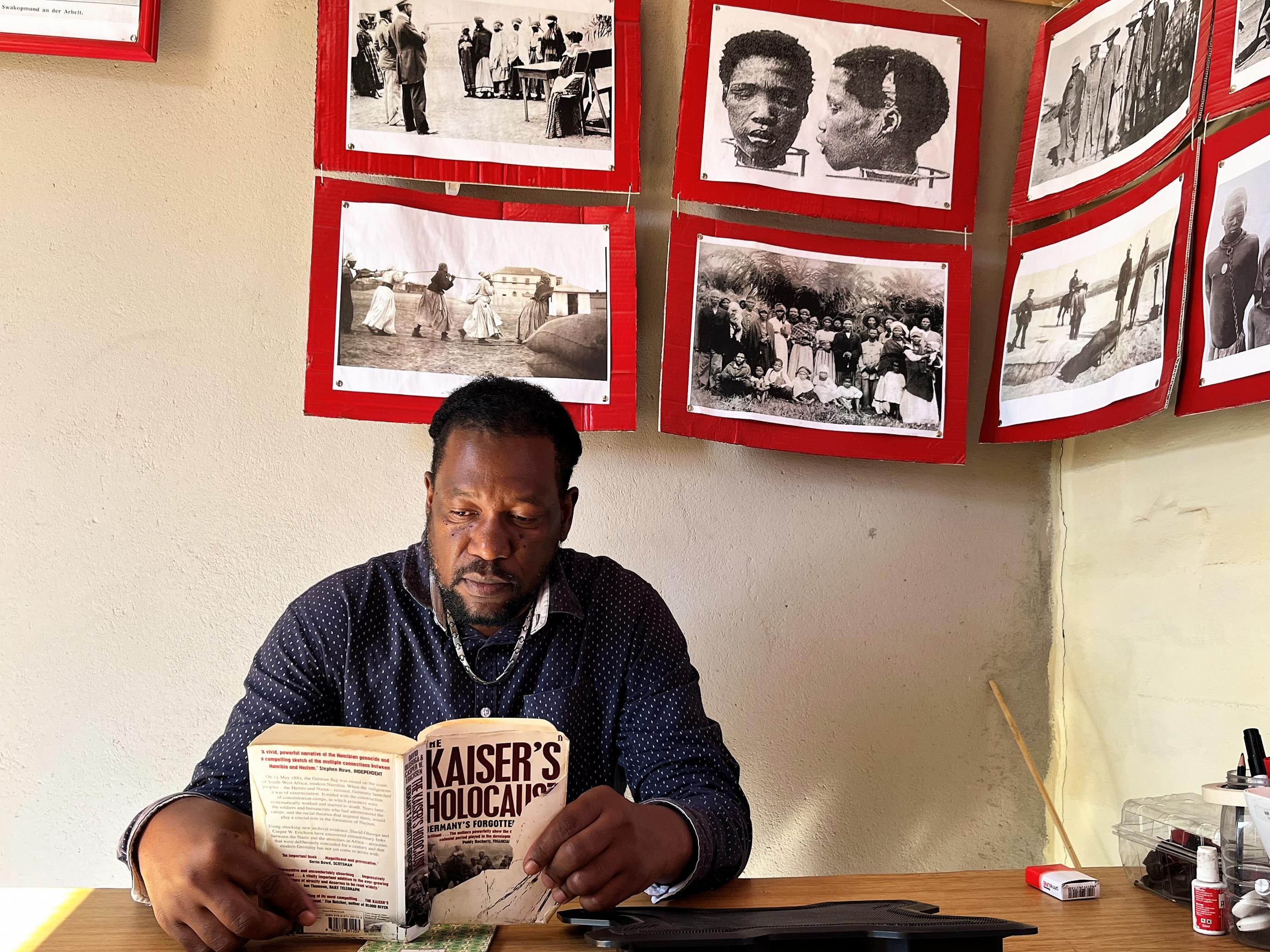 Der Mann sitzt an seinem Schreibtisch. Er liest im Buch „Kaisers Holocaust“. An der Wand hängen historische Fotos der Konzentrationslager in Swakopmund.